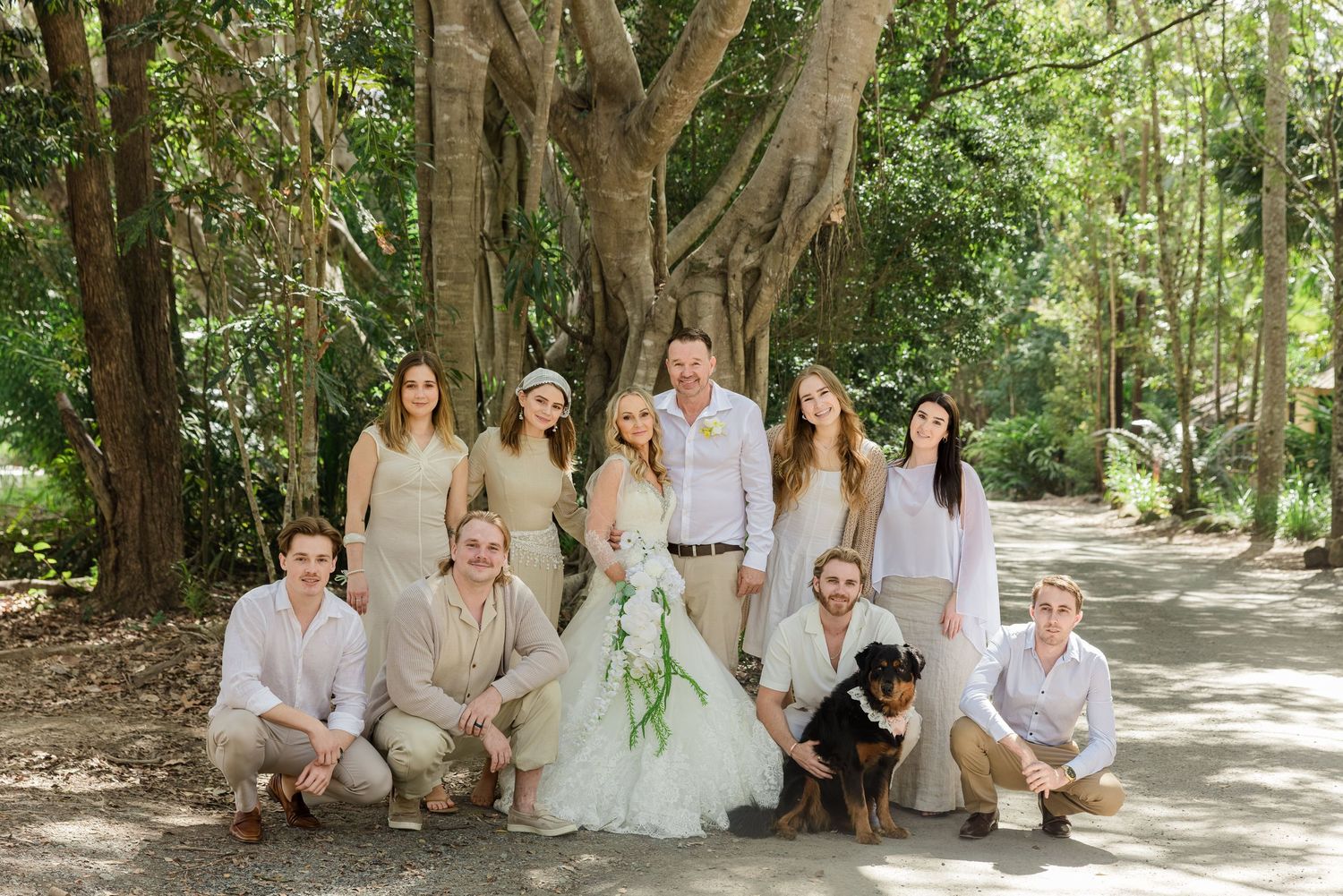 Wedding party poses outdoors under large trees, bride in white gown surrounded by guests in neutral tones with a black dog.