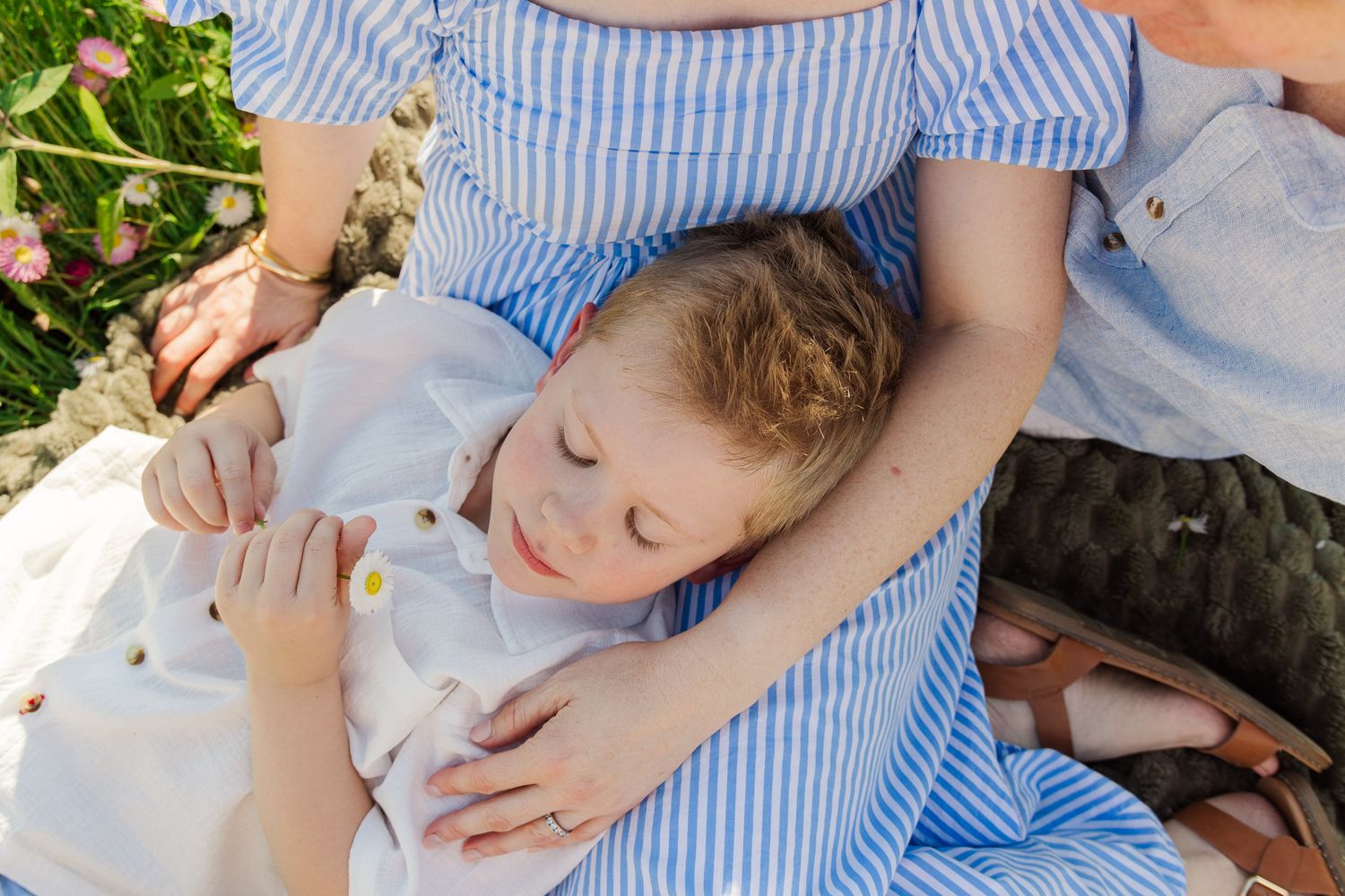 A child rests peacefully in a mother's arms outdoors, both wearing blue striped outfits among pink flowers.