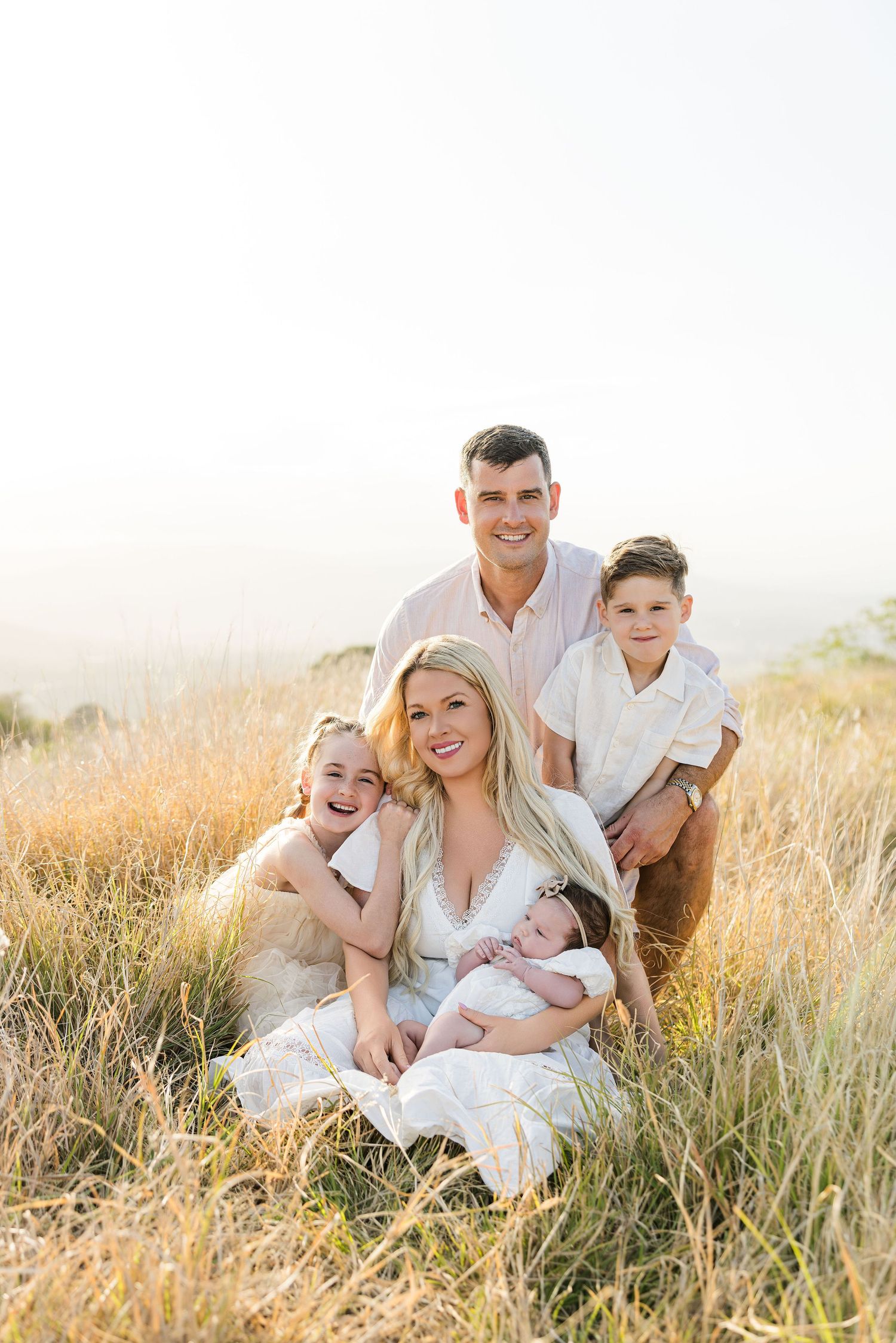 Family of five posing in golden field, mother holding newborn, father and two children smiling outdoors.