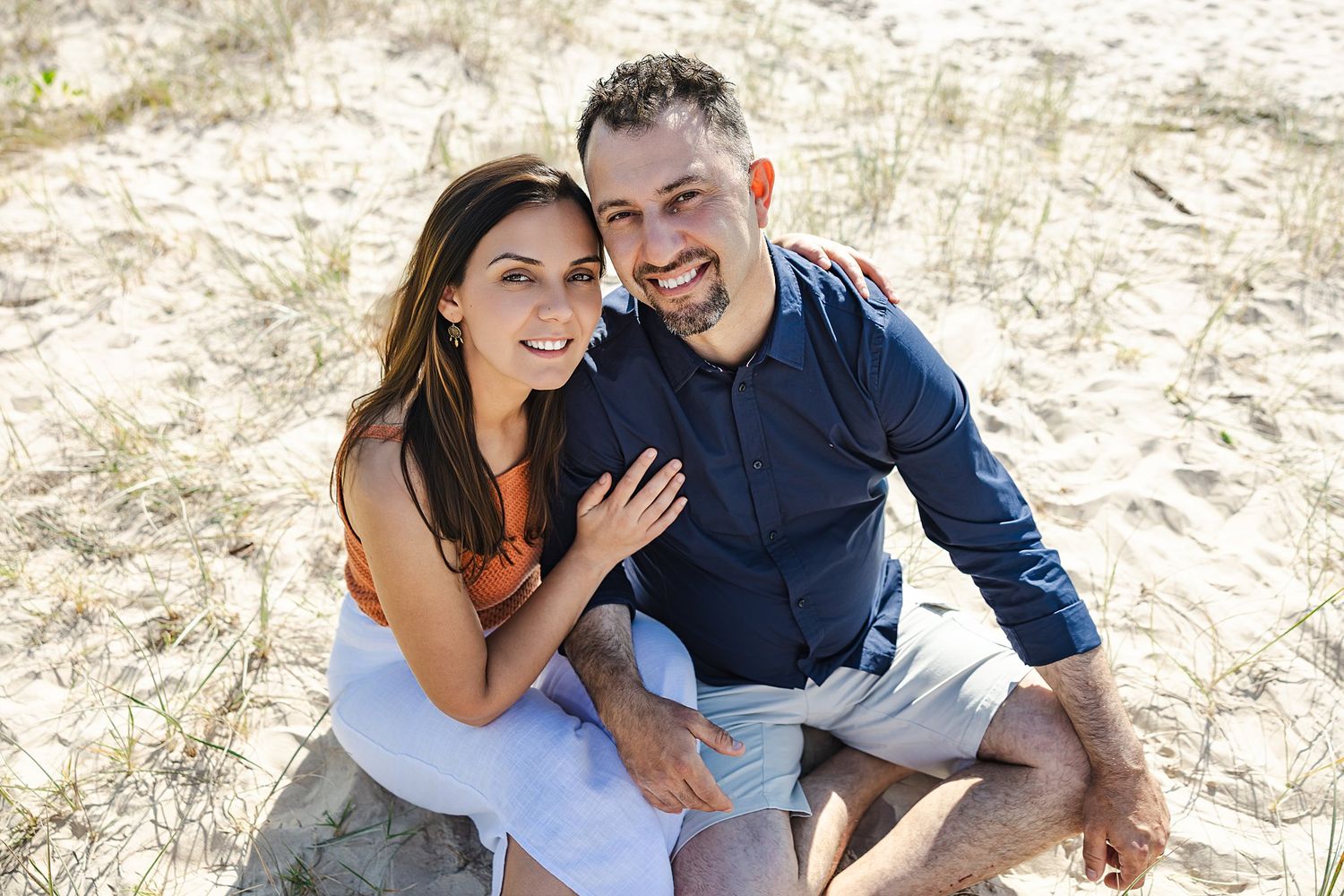 A couple sits together on a sandy beach, smiling warmly in casual attire during a sunny day.
