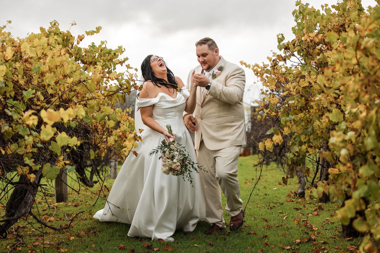 Joyful bride and groom laughing together among autumn vineyard rows, bride holding bouquet, groom in tan suit.