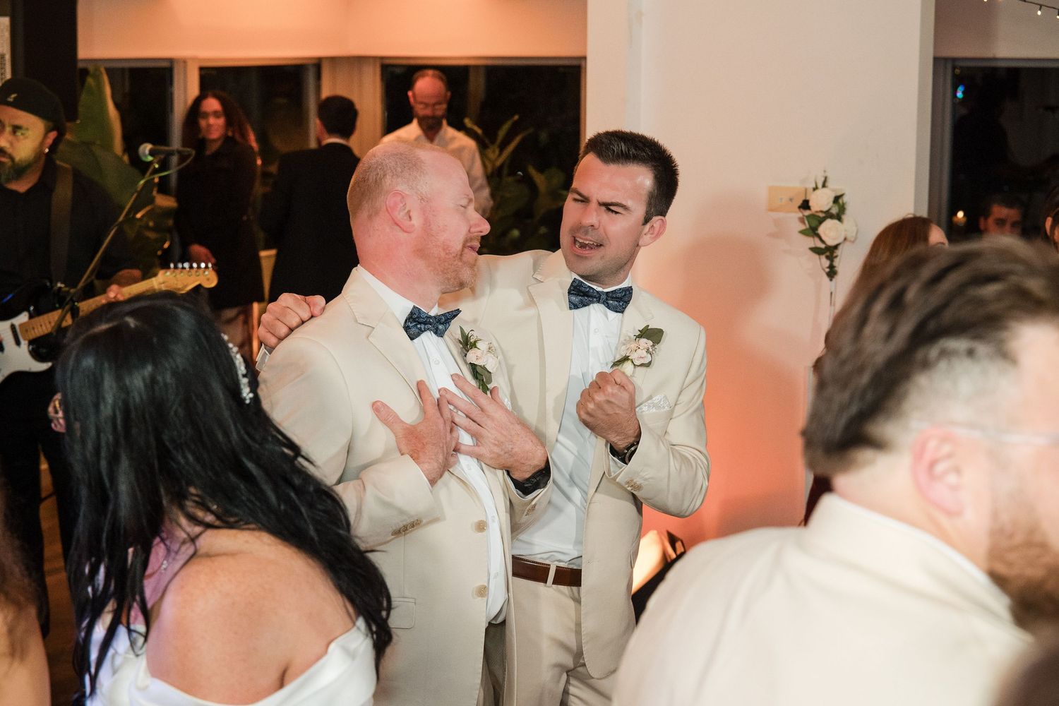 Two grooms in beige suits with bow ties share a joyful moment on the wedding dance floor.