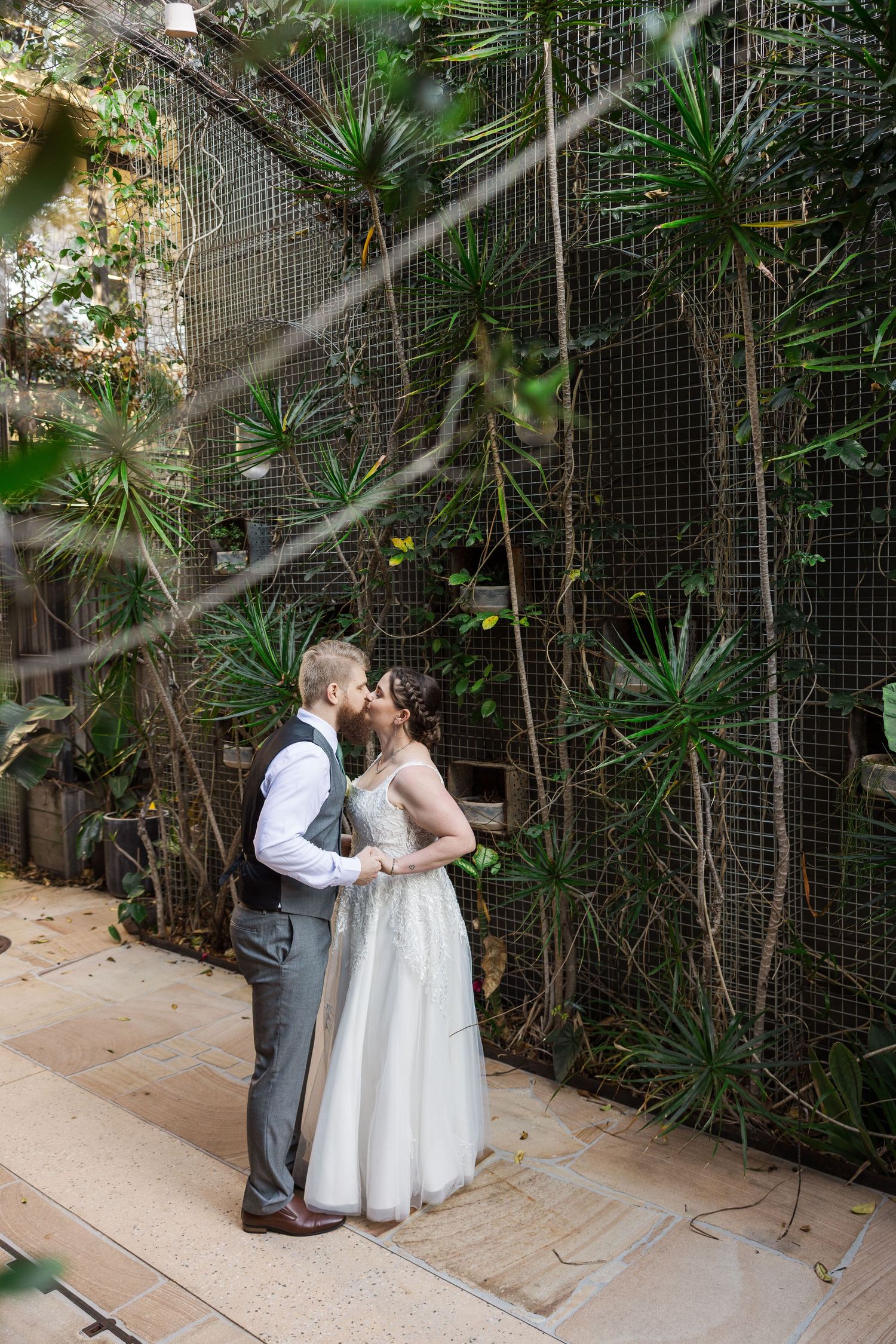 Wedding couple shares a kiss in a botanical garden with exotic plants and a metal trellis backdrop.