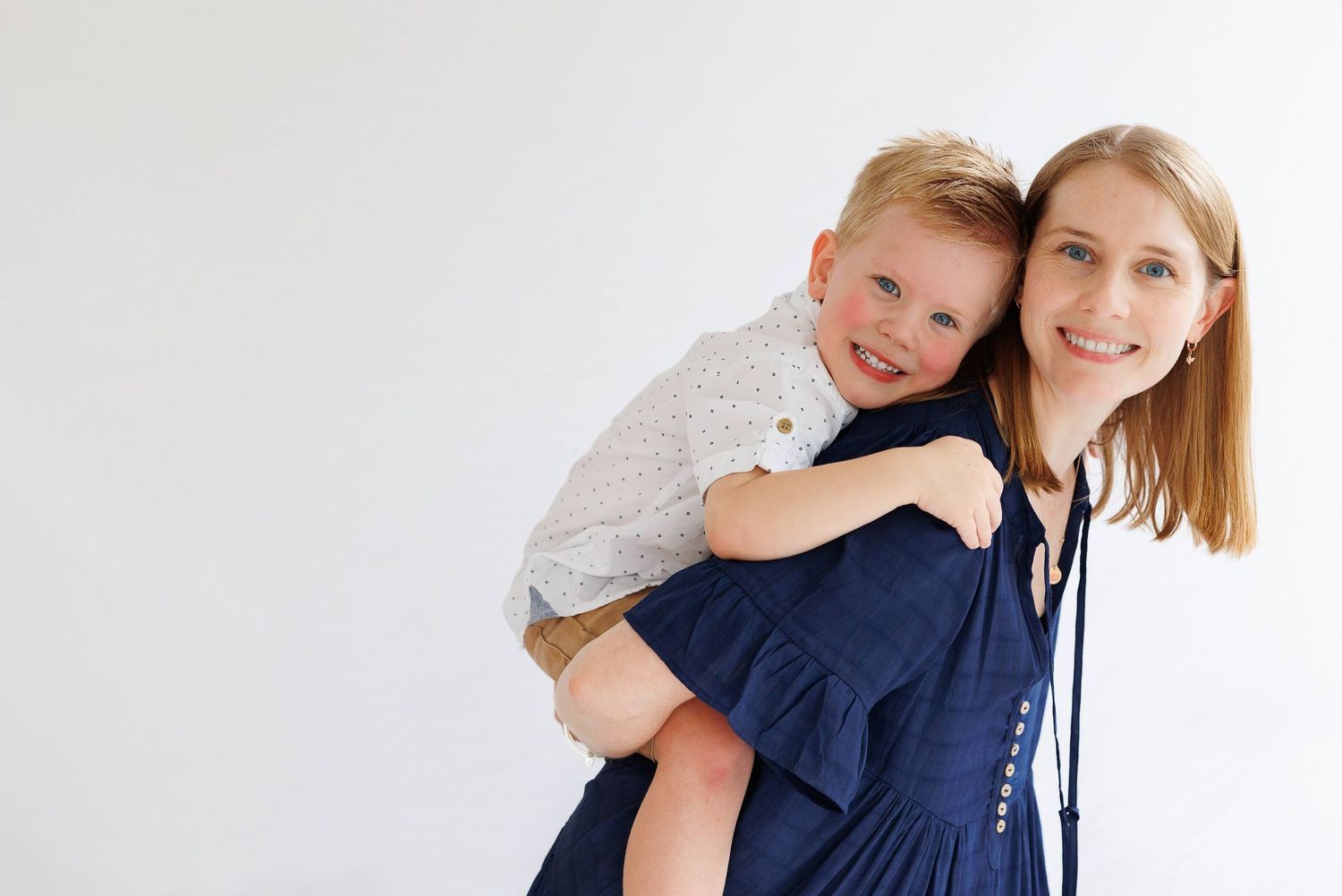 A smiling woman in a navy dress giving a happy young blonde boy a piggyback ride against a white background.