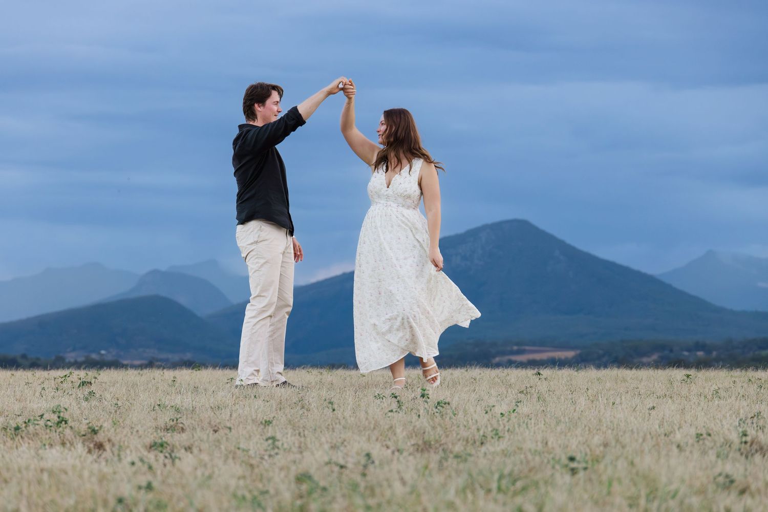 Couple dancing in a field with mountains and blue sky backdrop, man in black shirt twirling woman in white dress.