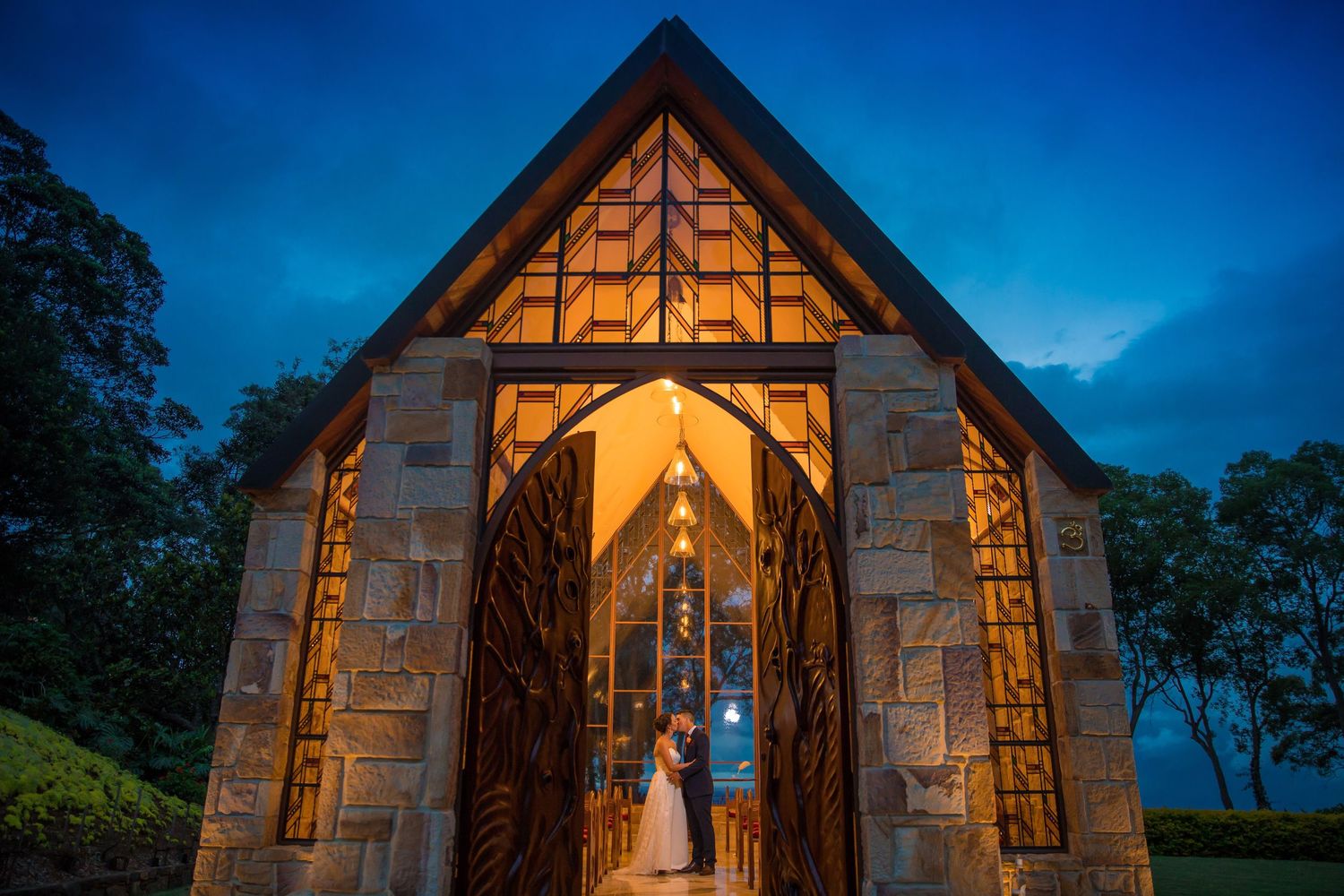 Couple standing in glowing stone chapel doorway at dusk, warm light illuminating wedding ceremony in scenic outdoor setting.
