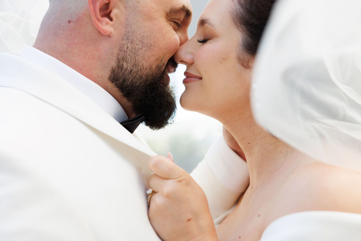 A bride and groom share a romantic almost-kiss moment on their wedding day, draped in a soft white veil.