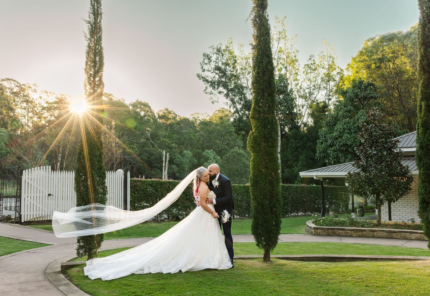 Bride and groom share romantic kiss at sunset in lush garden with tall cypress trees and flowing wedding veil.
