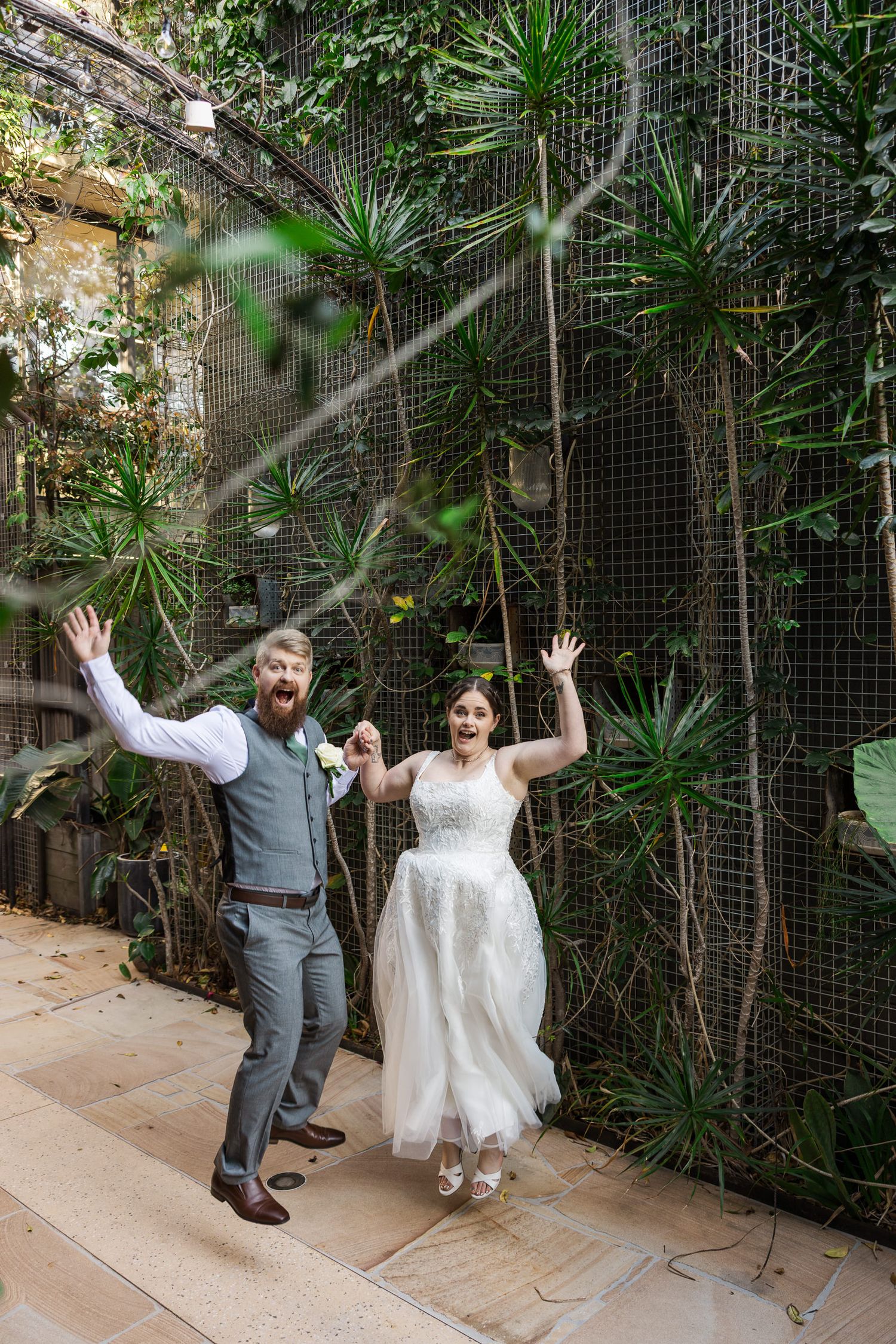 Joyful newlyweds celebrate in lush botanical garden, bride in white gown and groom in gray vest raising hands.
