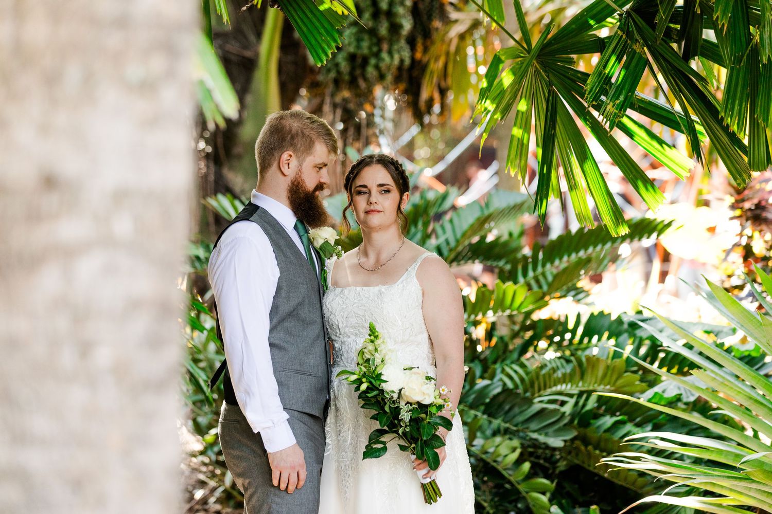 Bride and groom sharing an intimate moment surrounded by tropical palm fronds in a lush garden setting.