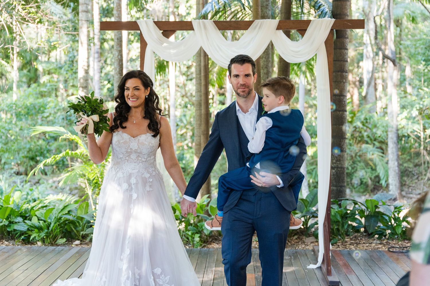 A wedding couple walks through a lush garden pathway with trees creating a natural archway overhead.