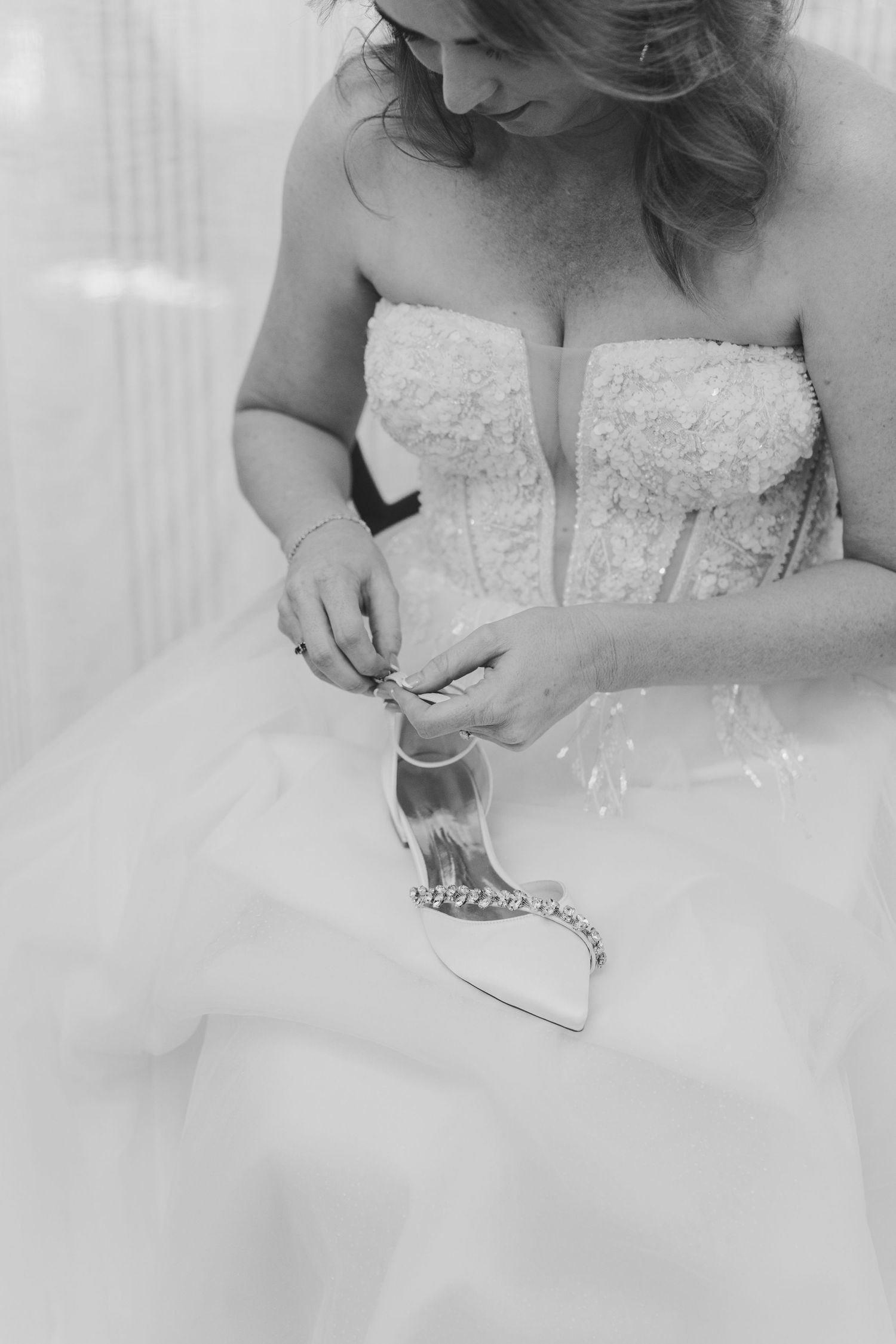 Black and white photo of a bride in a strapless lace wedding gown fastening her elegant bridal shoe.