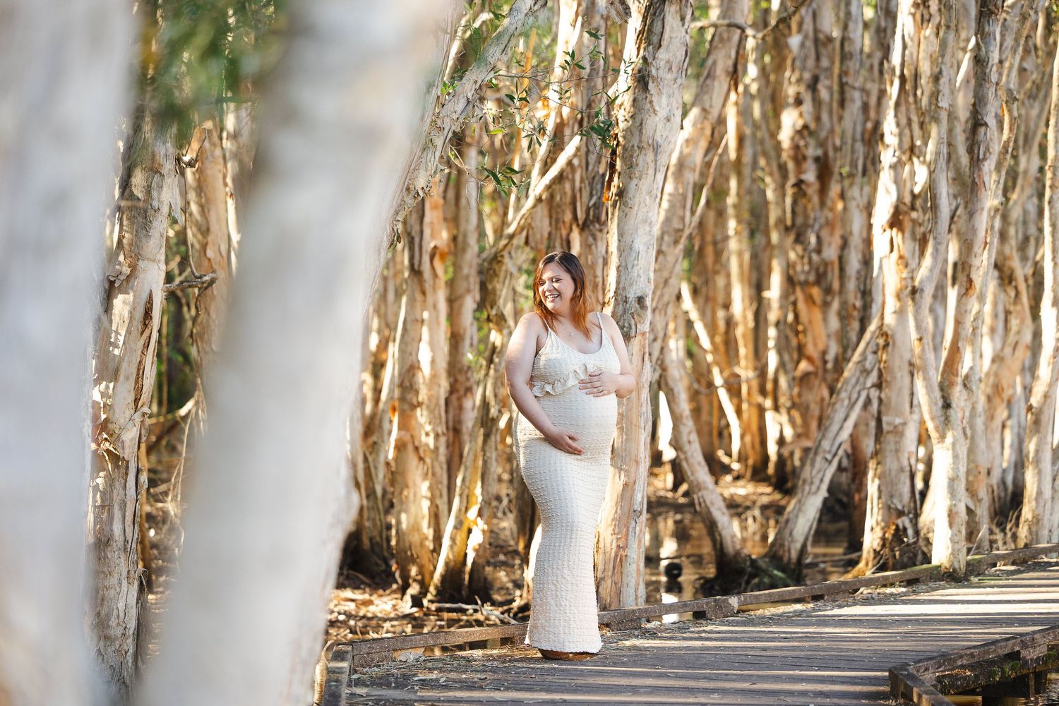 A figure in a flowing white dress stands in a scenic path lined with paperbark trees.