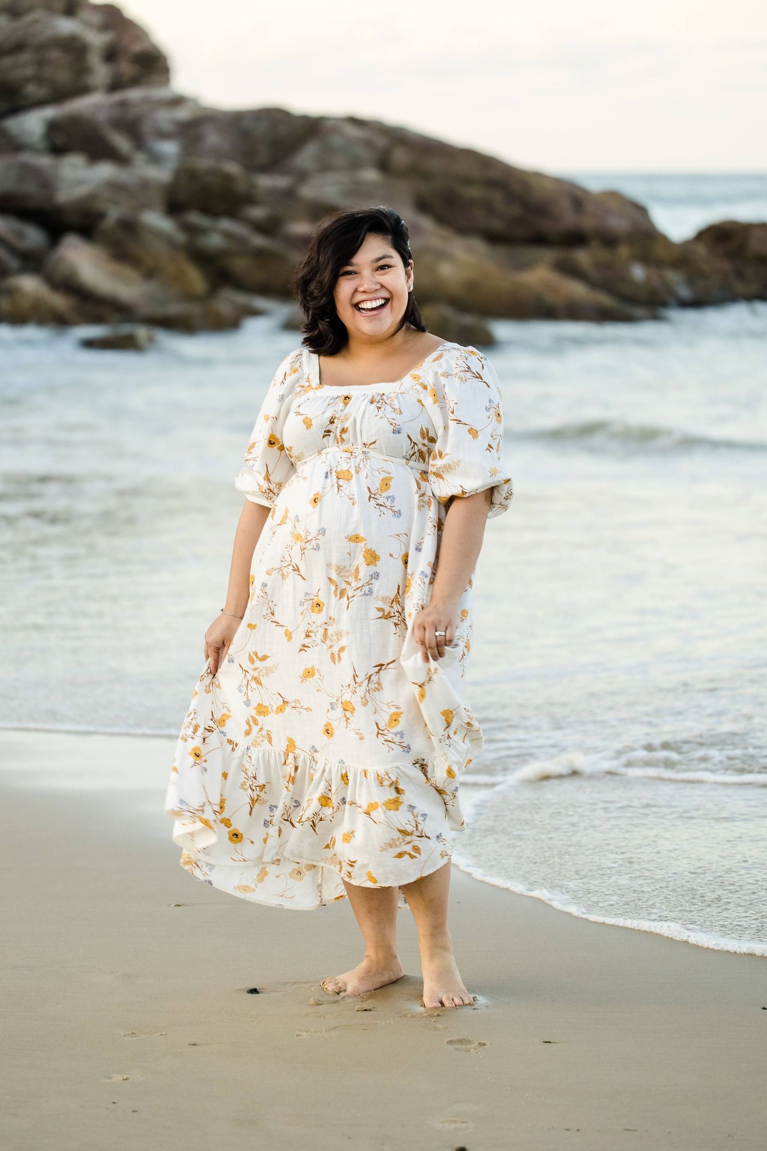 Someone in a floral white summer dress stands barefoot on a beach with rocky cliffs and ocean waves in the background.