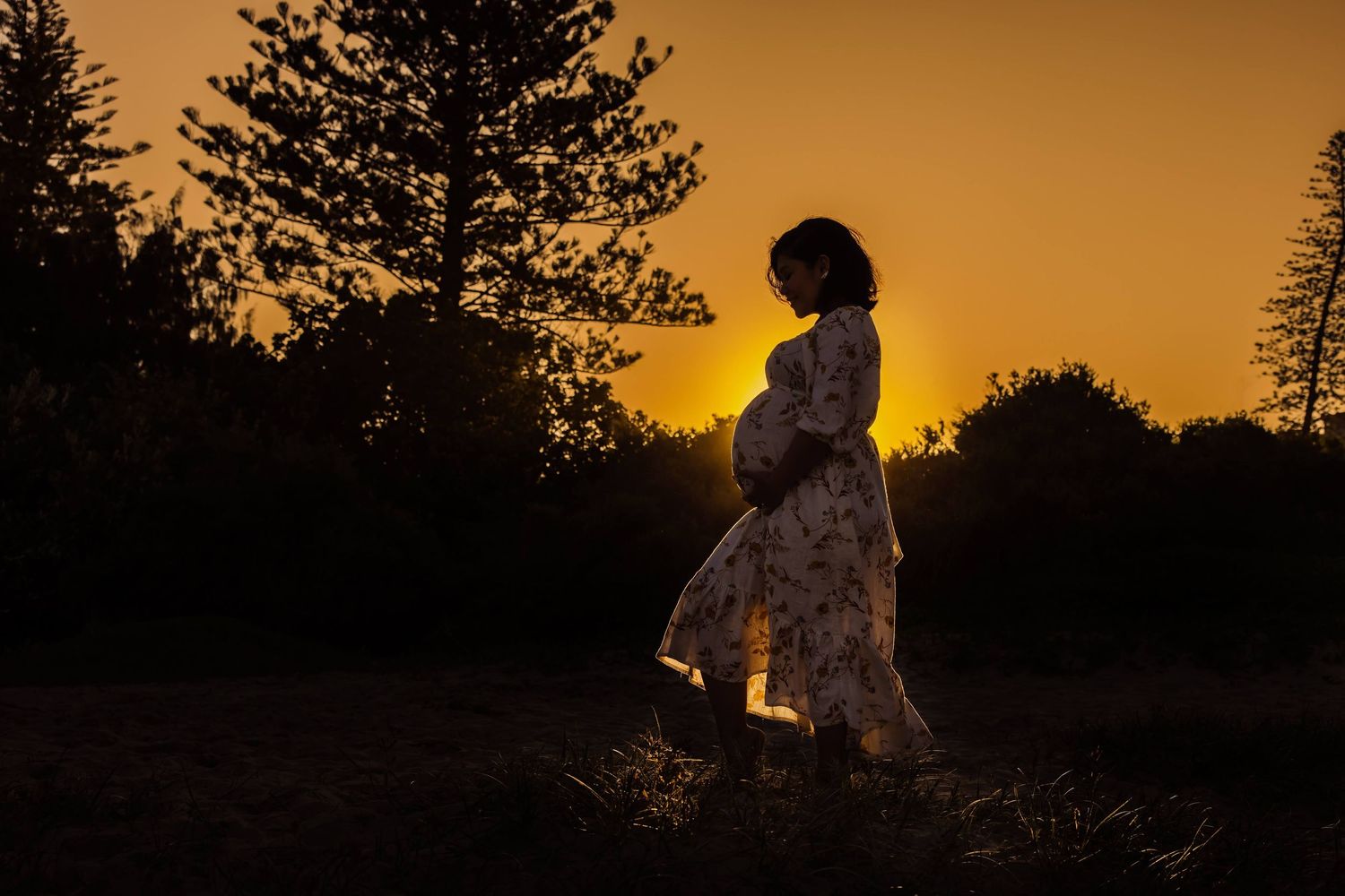A silhouette against a golden sunset stands in a flowy dress with pine trees in the background.