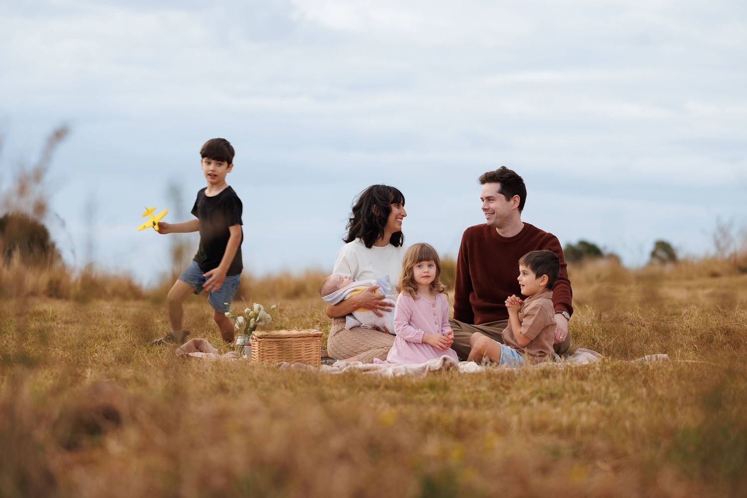 A family enjoys a summer picnic together in a golden field during sunset while someone plays frisbee in the background.