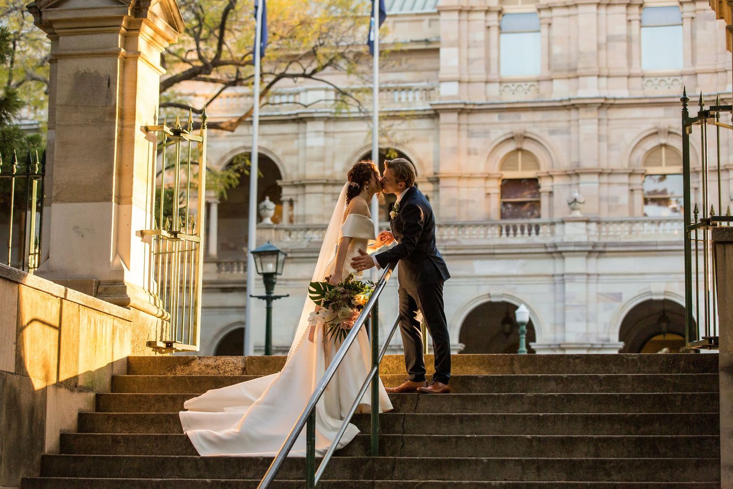 Elegant wedding couple poses on ornate stone staircase with warm sunlight illuminating the historic architecture.