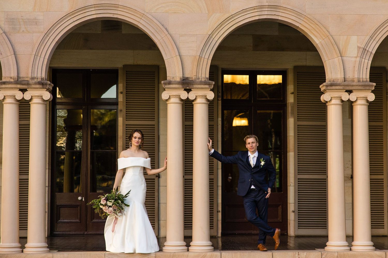 A wedding couple stands in an elegant stone archway colonnade with ornate architectural details and warm lighting.