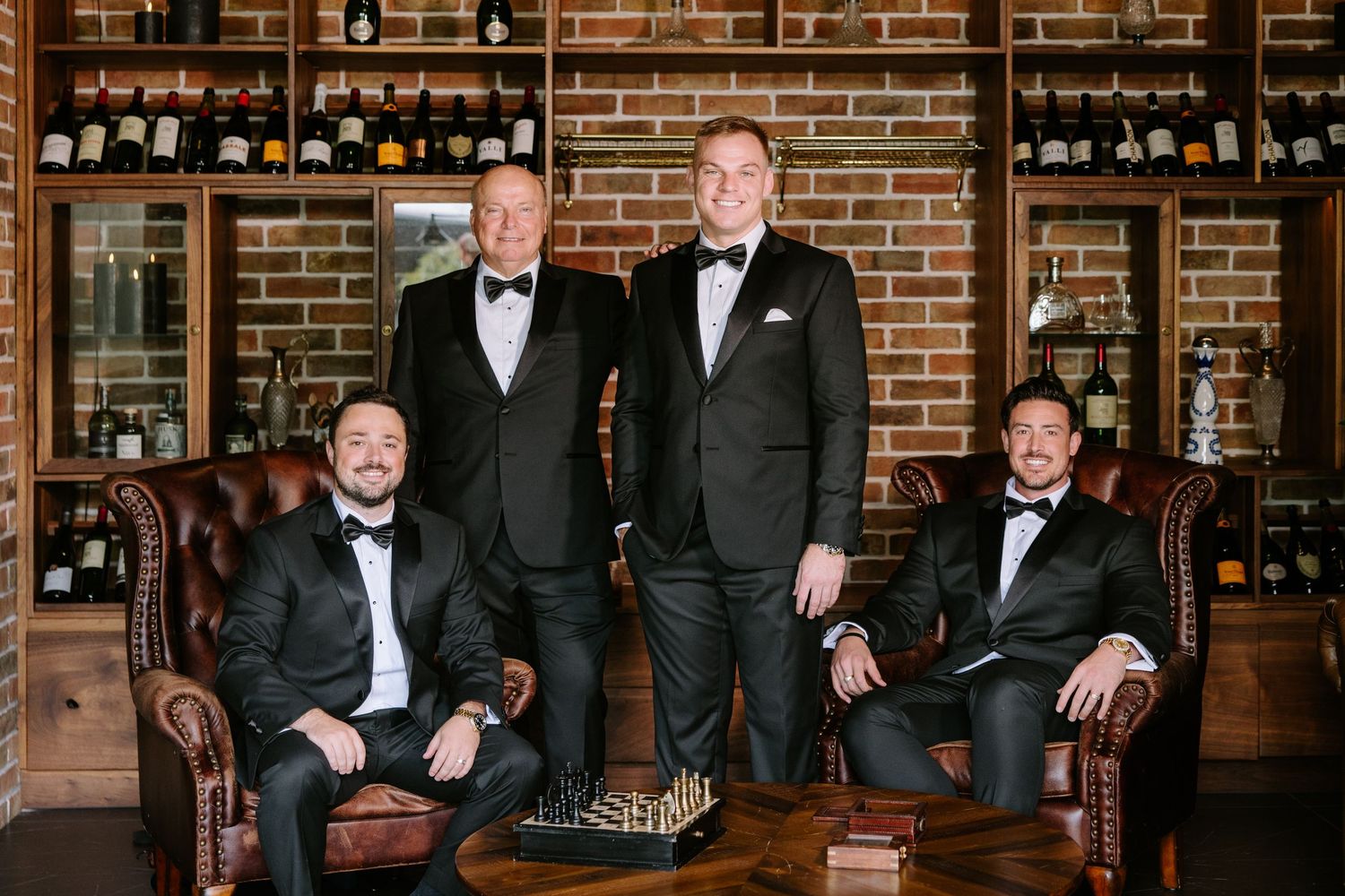Groomsmen in black tuxedos pose together in a rustic wine cellar with brick walls and wooden shelving.