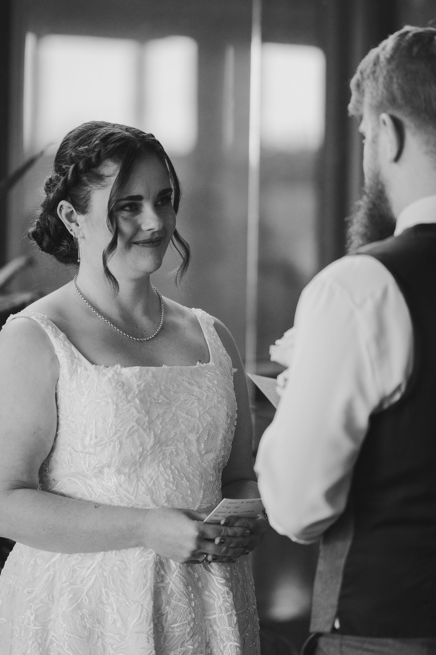 Black and white photo of a bride in lace dress exchanging vows with bearded groom during indoor wedding ceremony.