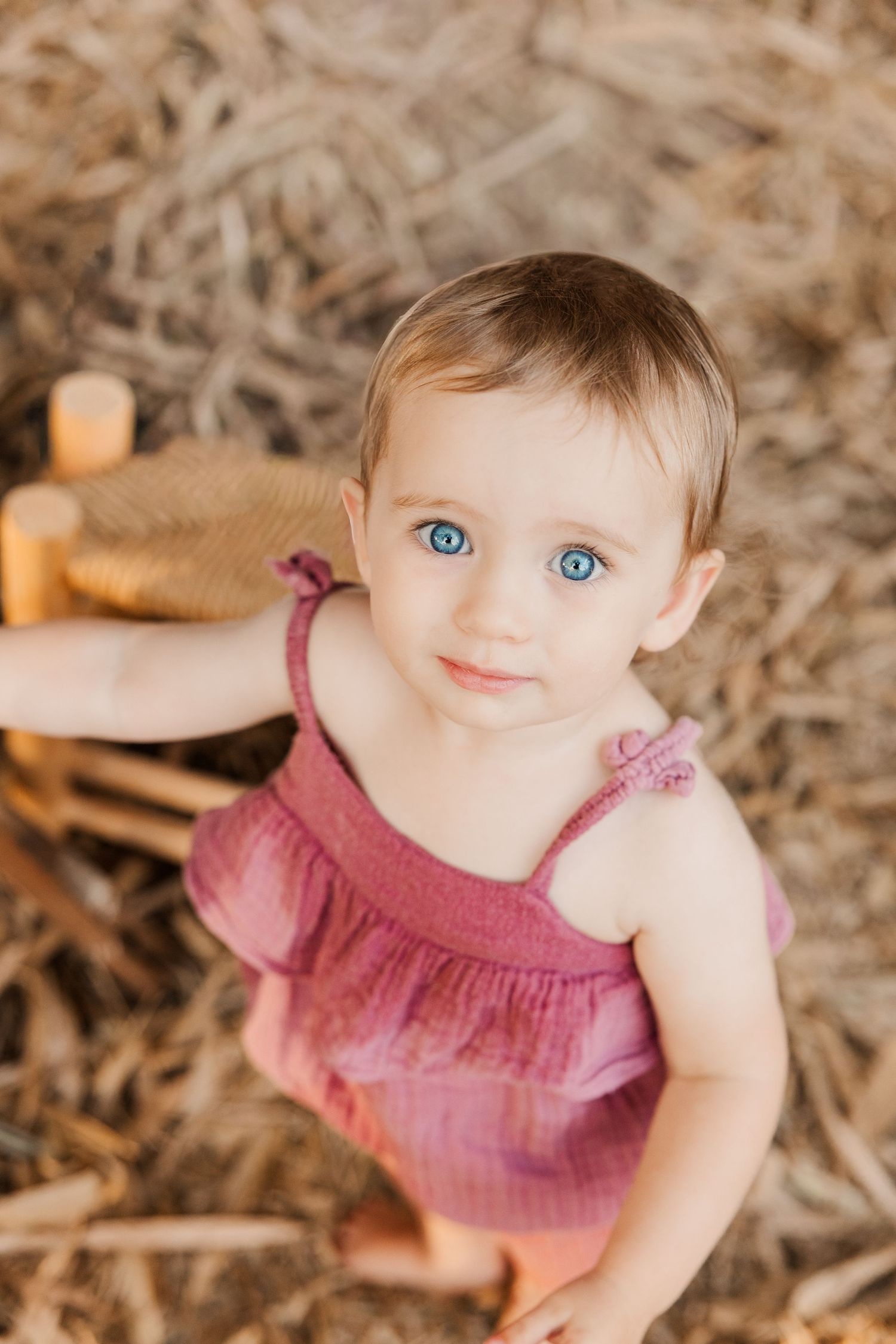 A child in a pink sleeveless dress sits outdoors on natural ground cover.