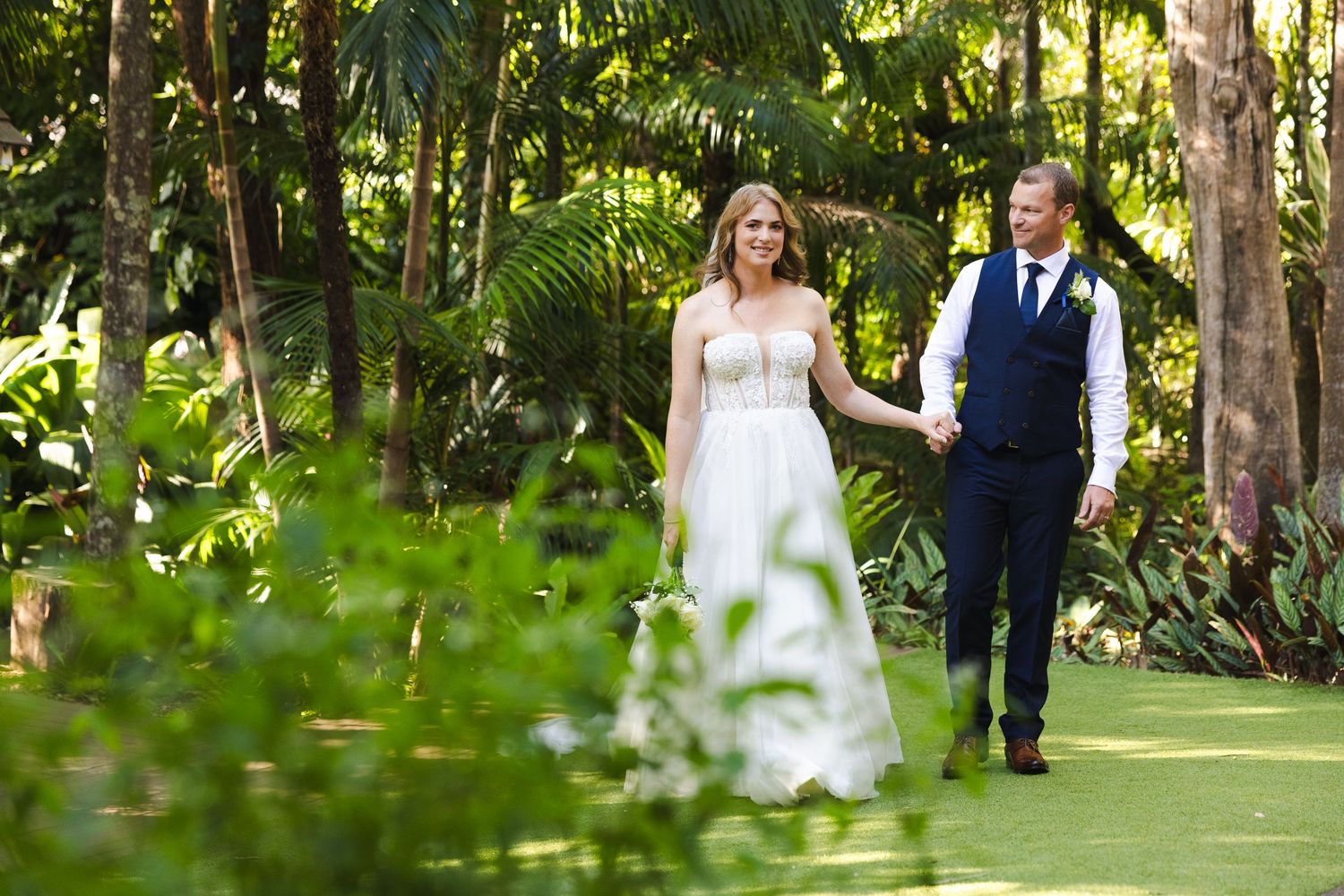 Newlyweds walk hand in hand through a lush tropical garden path surrounded by palm trees.