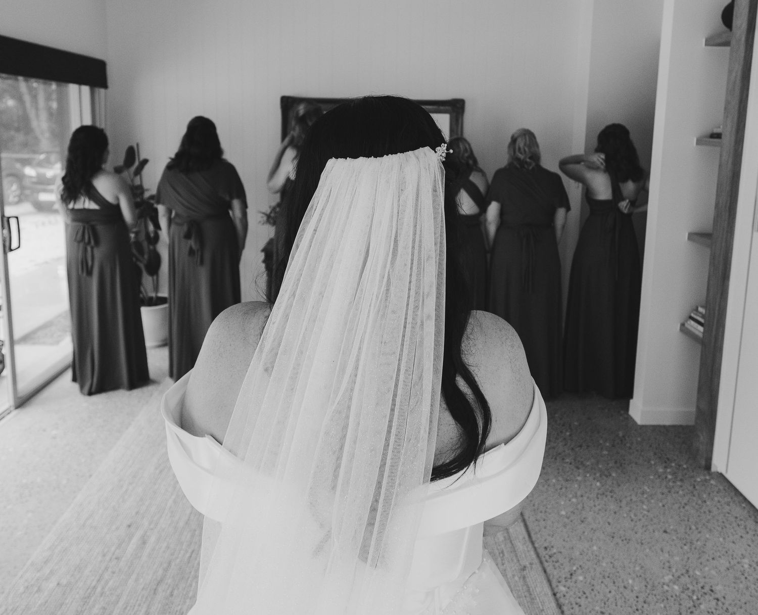 Black and white photo of a bride in a veil with bridesmaids reflected in a mirror during wedding preparations.