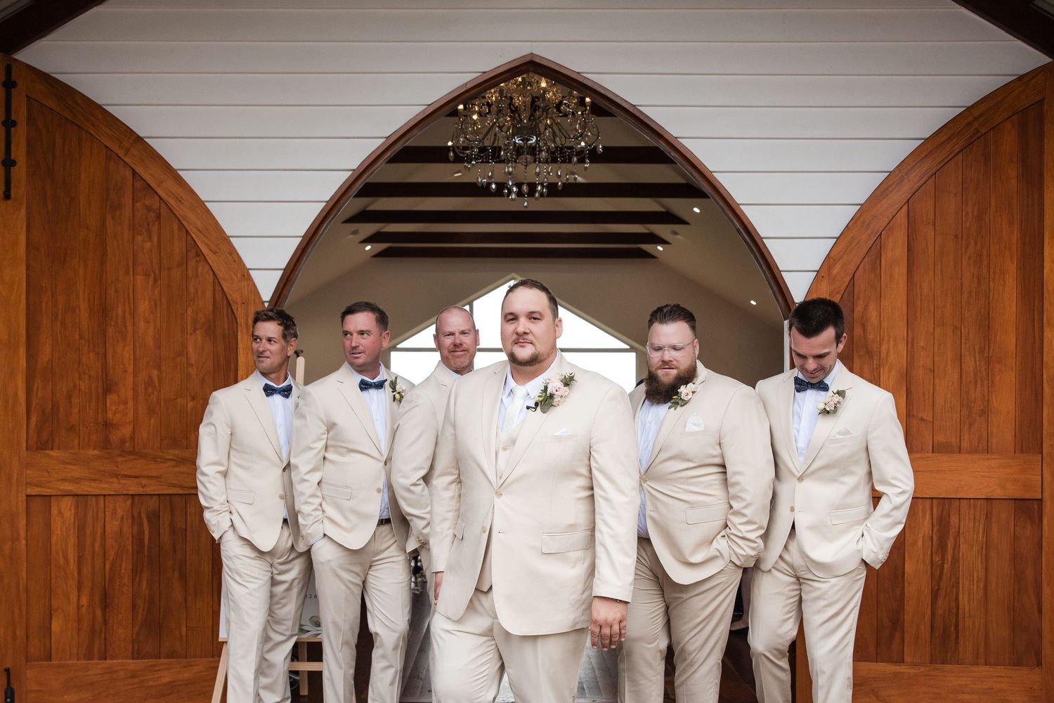 Groom in ivory suit poses with groomsmen in tan suits and bow ties before wooden chapel doors.