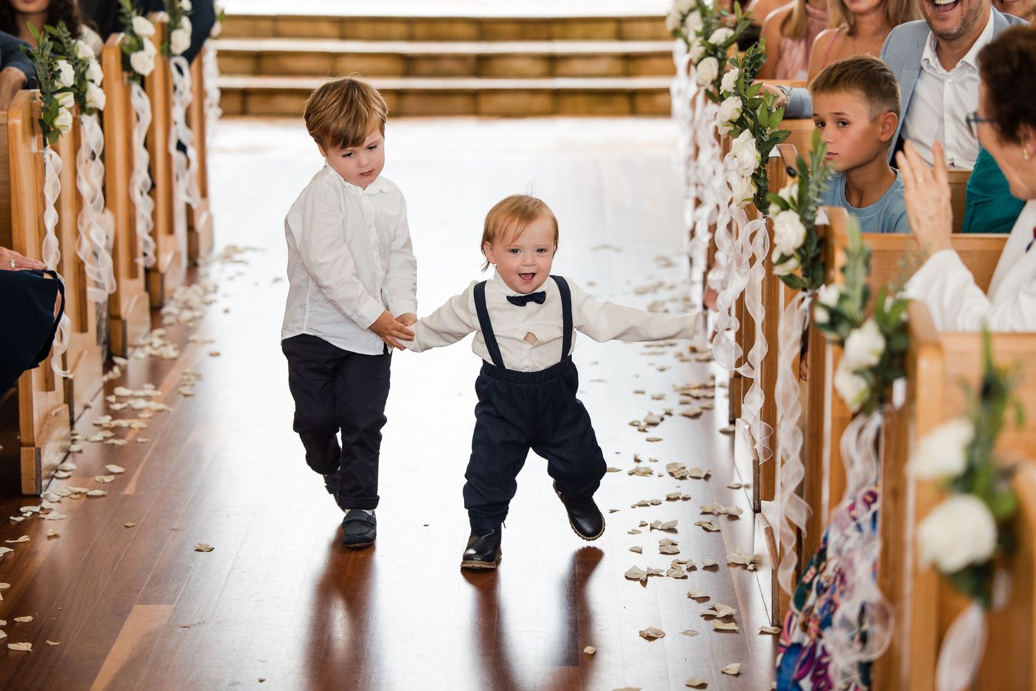 Two young ring bearers in bow ties run joyfully down the wedding aisle at a rustic barn ceremony.