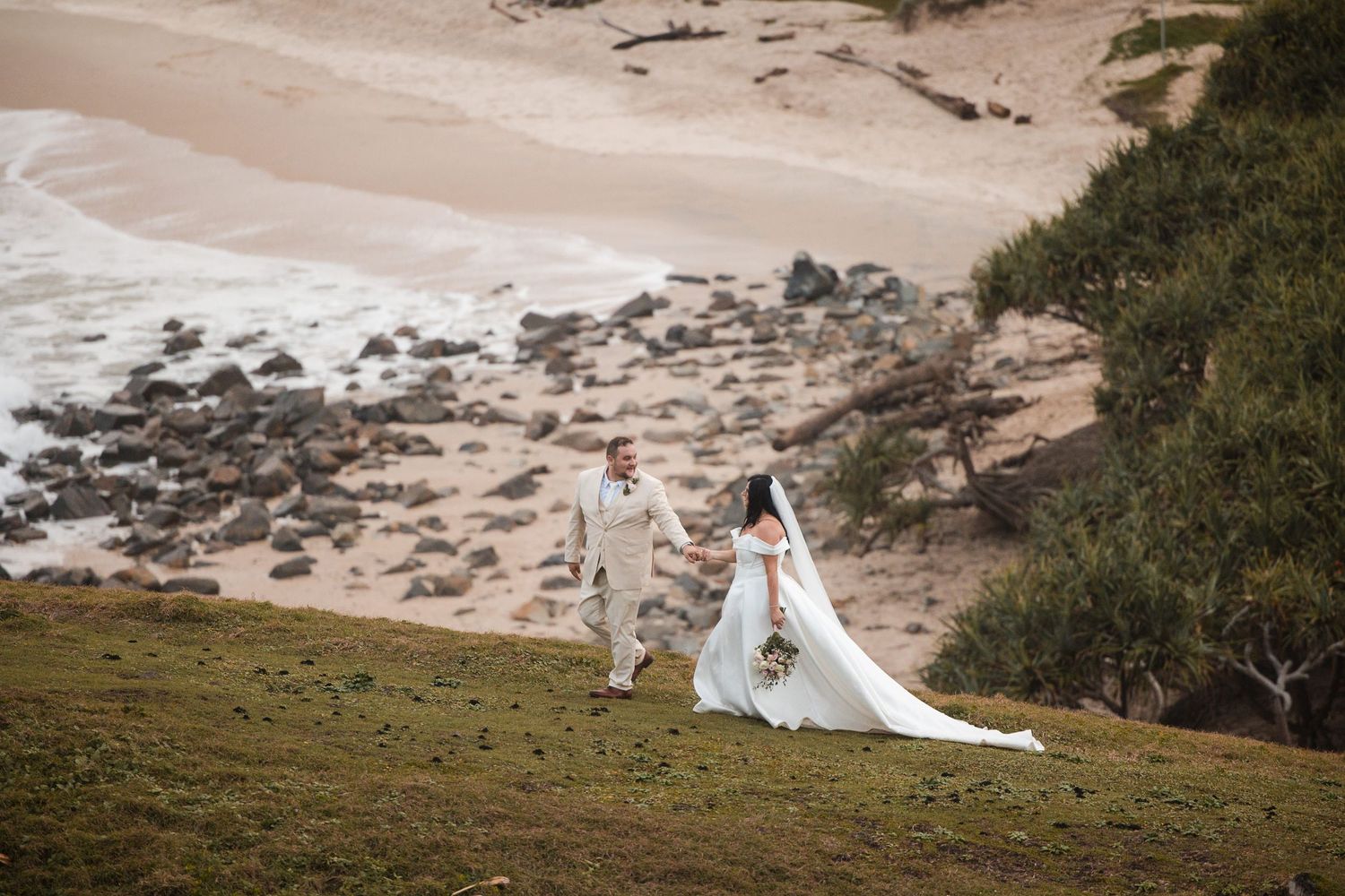 Bride and groom walk hand in hand on a grassy coastal cliff overlooking a rocky beach and ocean waves.
