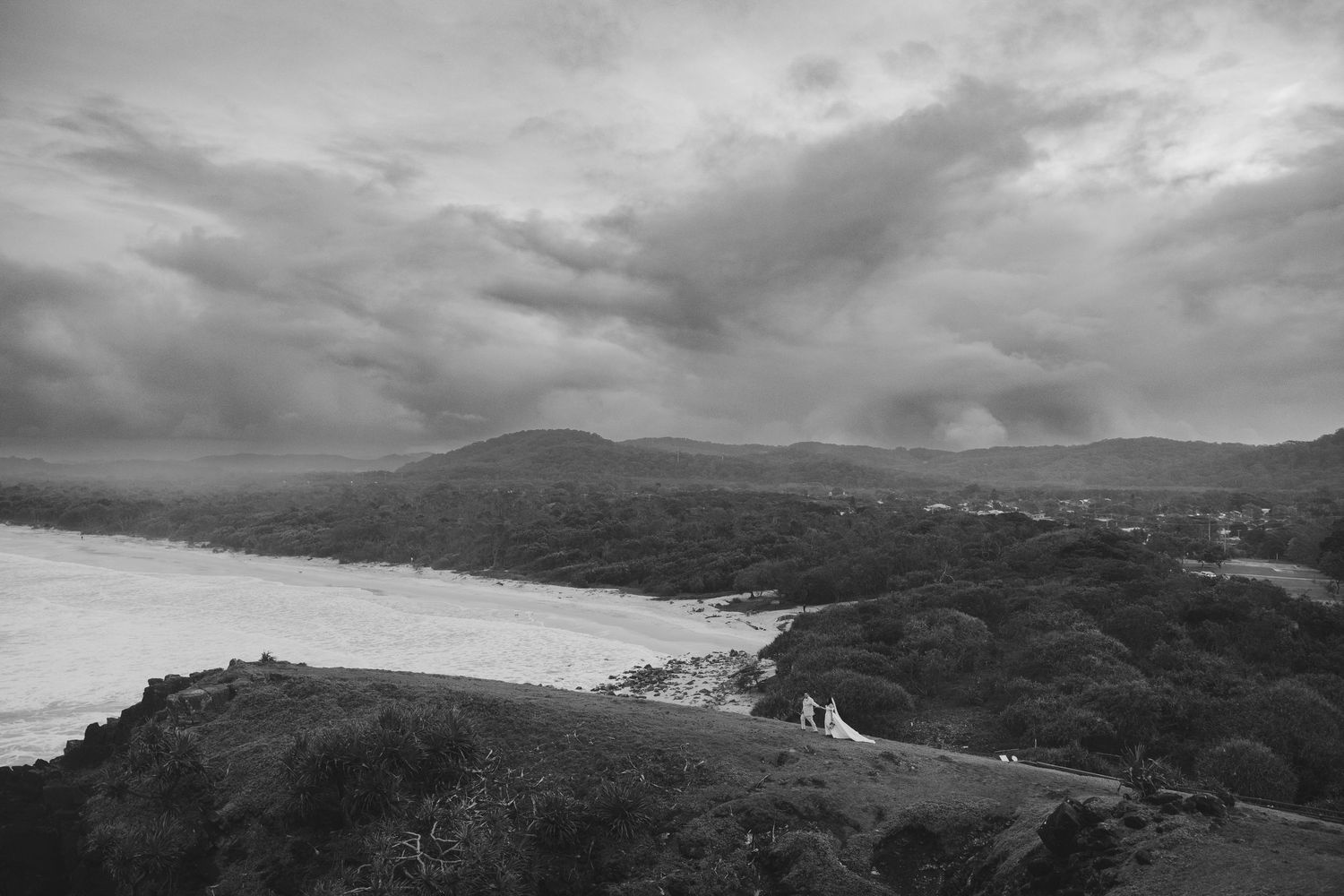 Black and white aerial view of a river winding through lush hills under dramatic cloudy skies with two figures on a path.