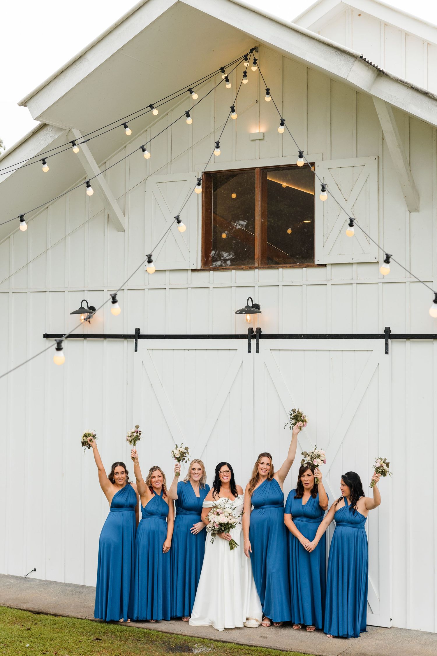 Bride and bridesmaids in blue dresses celebrating in front of a white barn with string lights.