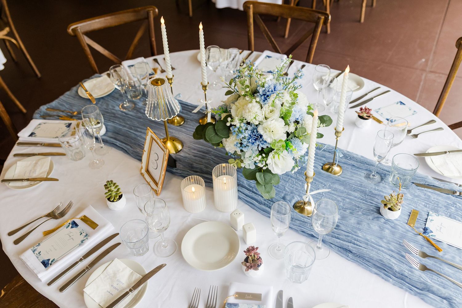 Elegant wedding reception table with blue hydrangea centerpiece, gold candlesticks, white linens, and blue table runner.