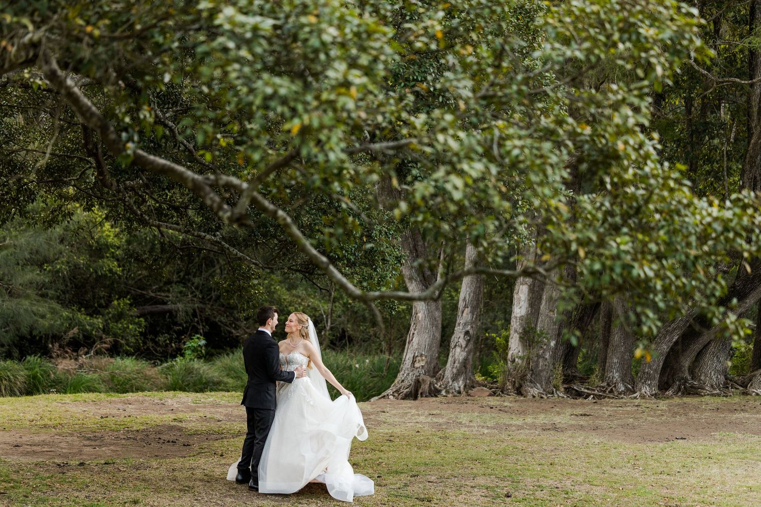 Bride and groom share a romantic moment in a lush green garden surrounded by tall trees on their wedding day.