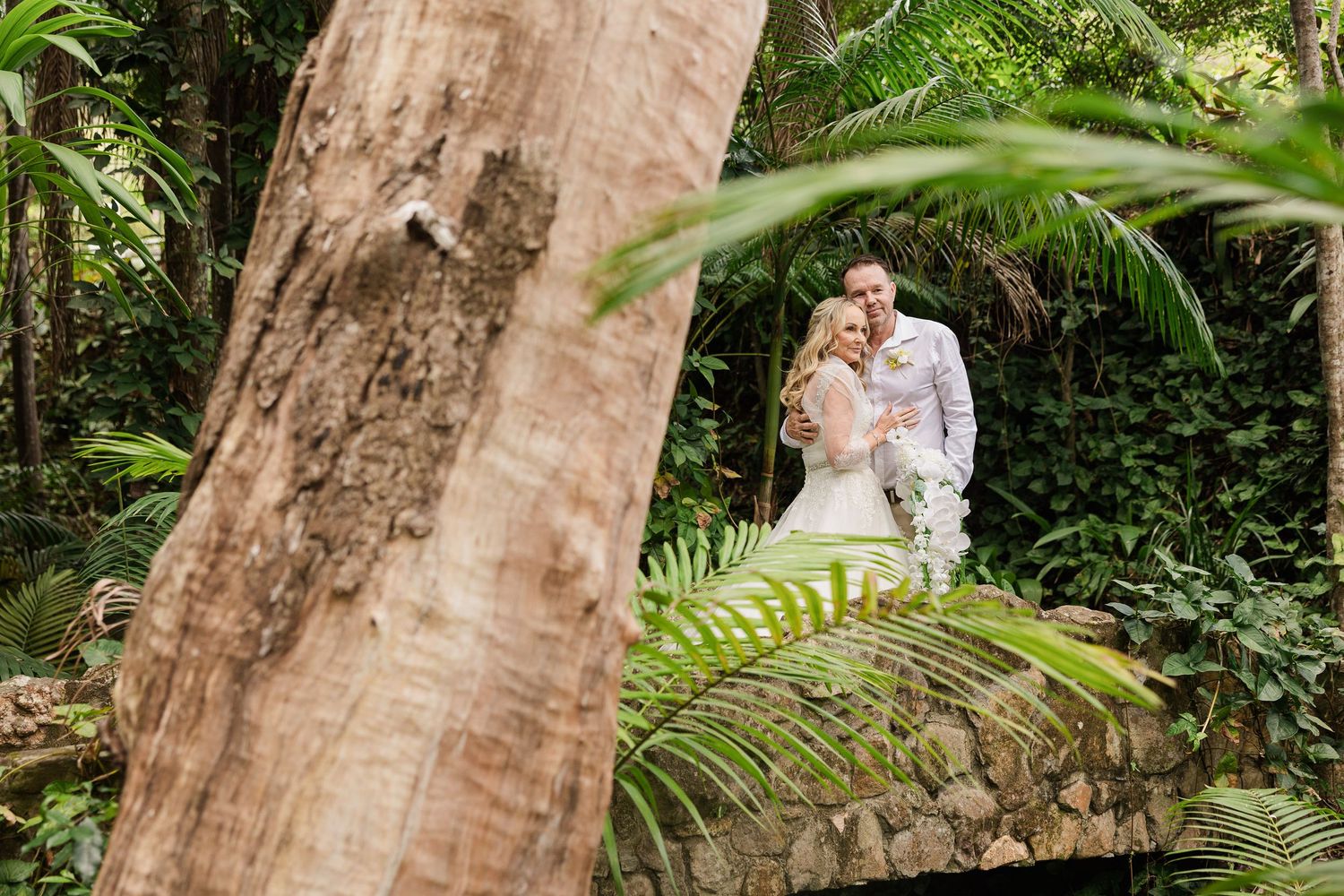 A couple in wedding attire embraces on a stone bridge surrounded by lush tropical greenery and palm trees.
