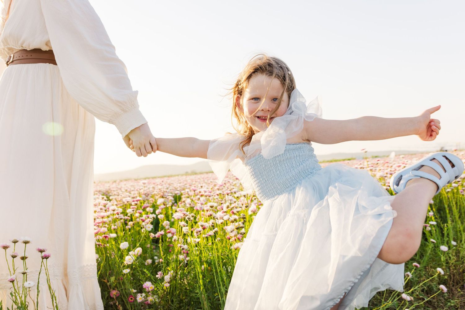 Young girl in white dress laughing joyfully in a flower field, holding hands with adult in white robe on sunny day.