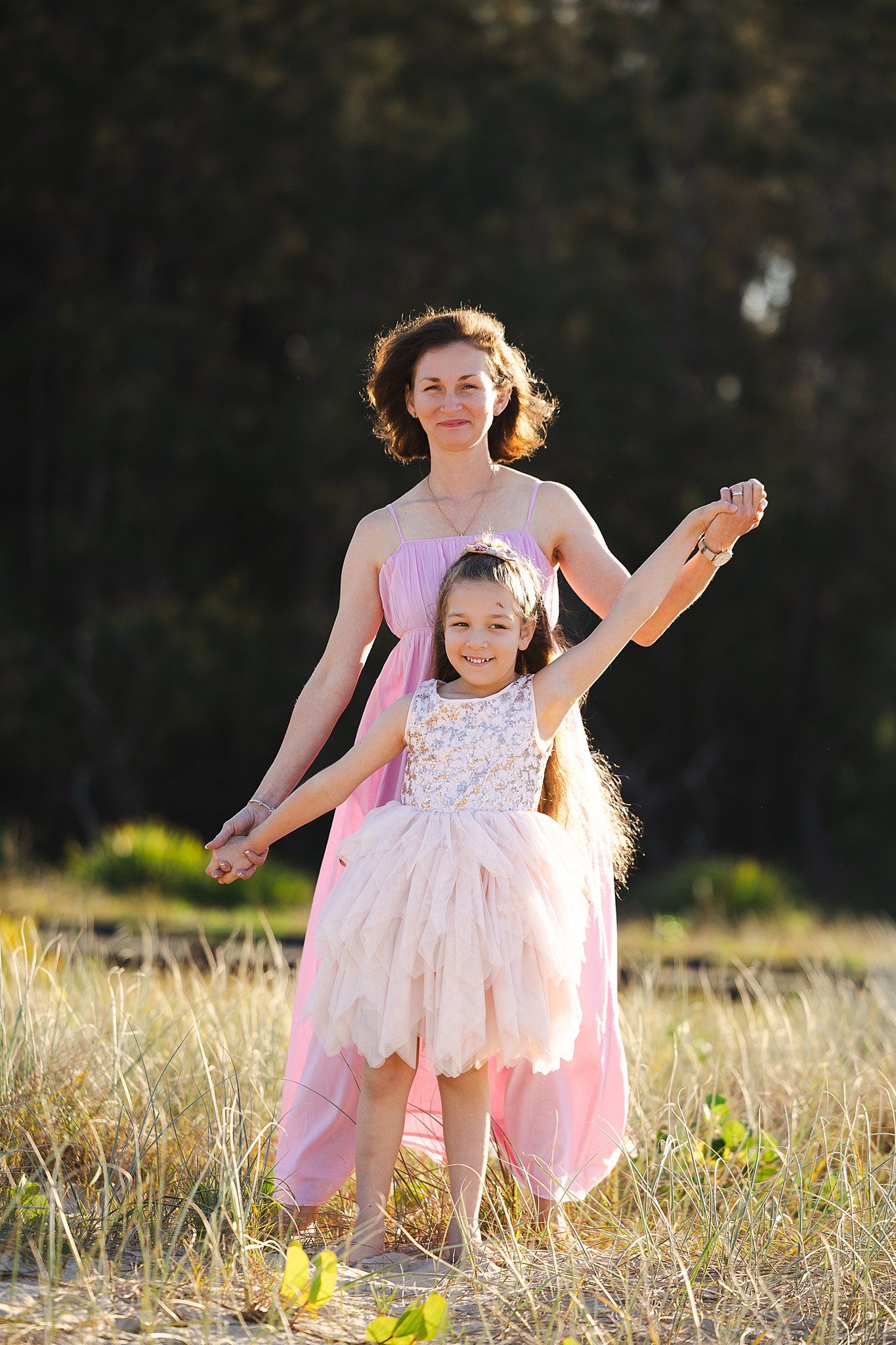 A parent and child wearing pink dresses dance together in a sunlit meadow at golden hour.