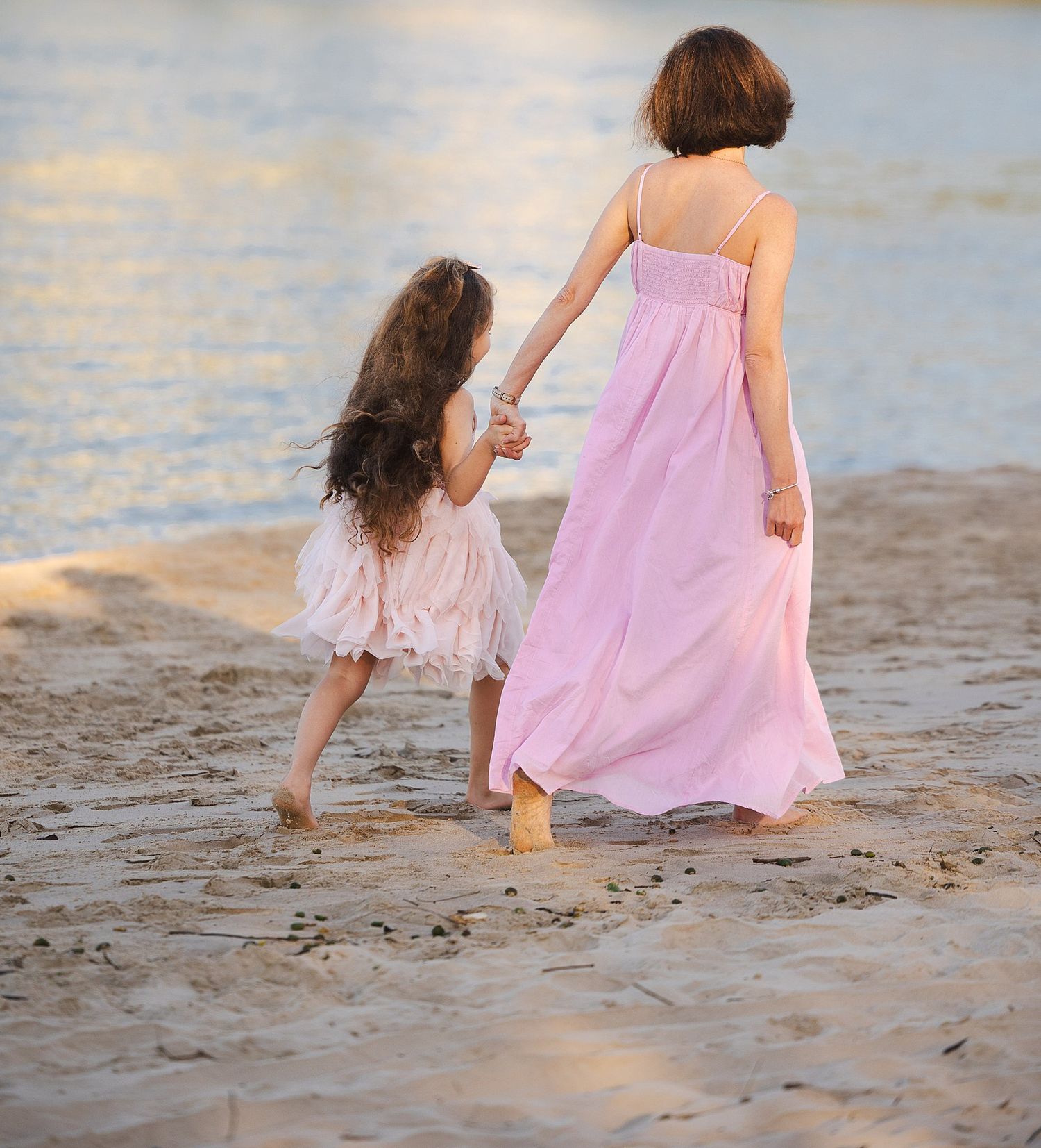 A sentimental beach scene of a parent and child in matching pink dresses walking hand in hand along the shoreline at sunset.