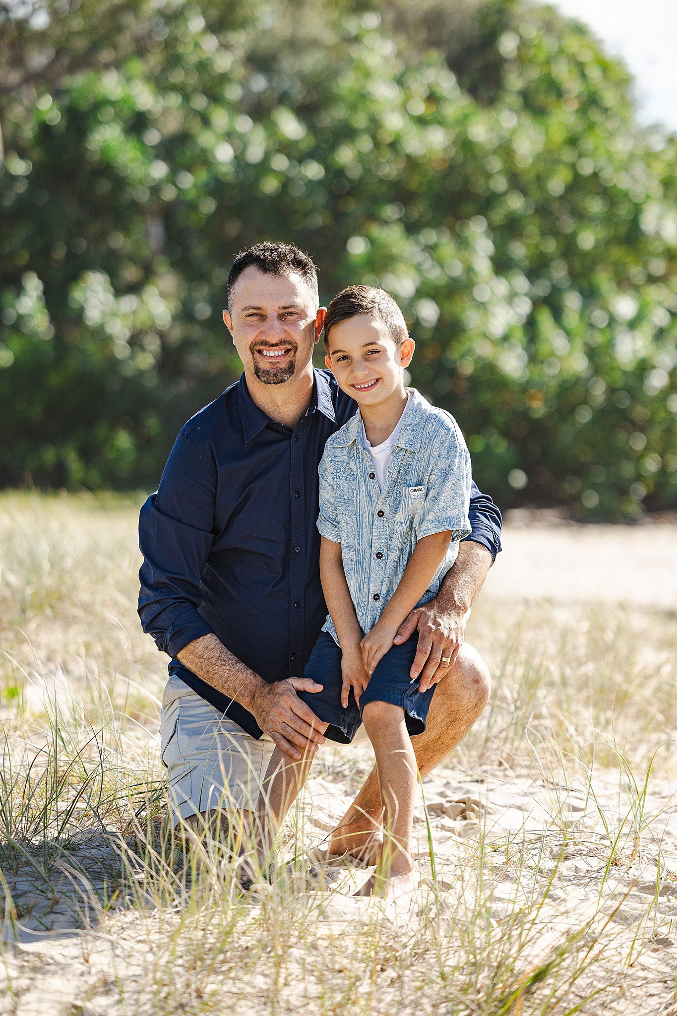 Two people pose together on sandy terrain with green vegetation in the background on a sunny day.