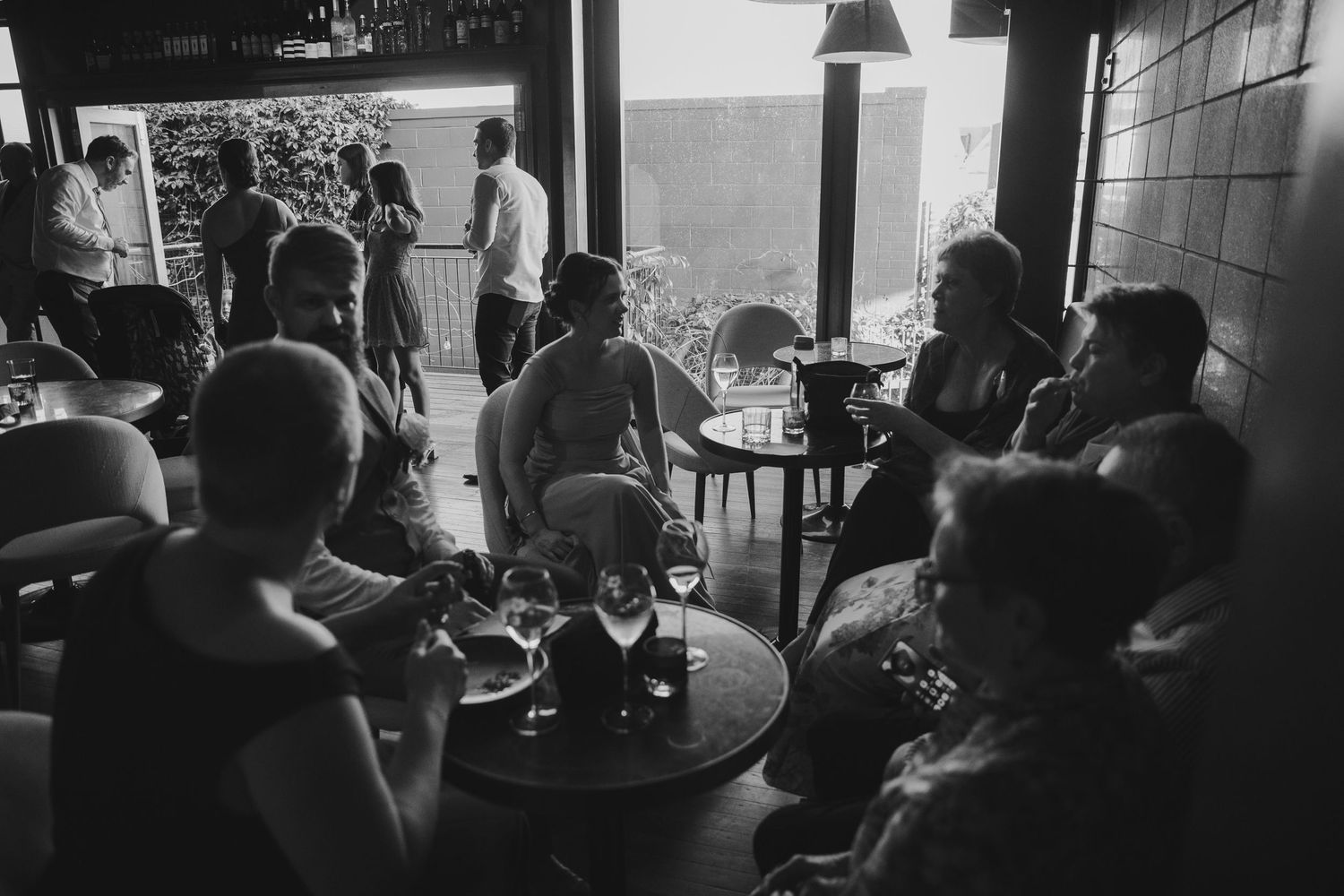 Black and white photo of guests socializing in a dimly lit bar or restaurant with wine glasses on tables.