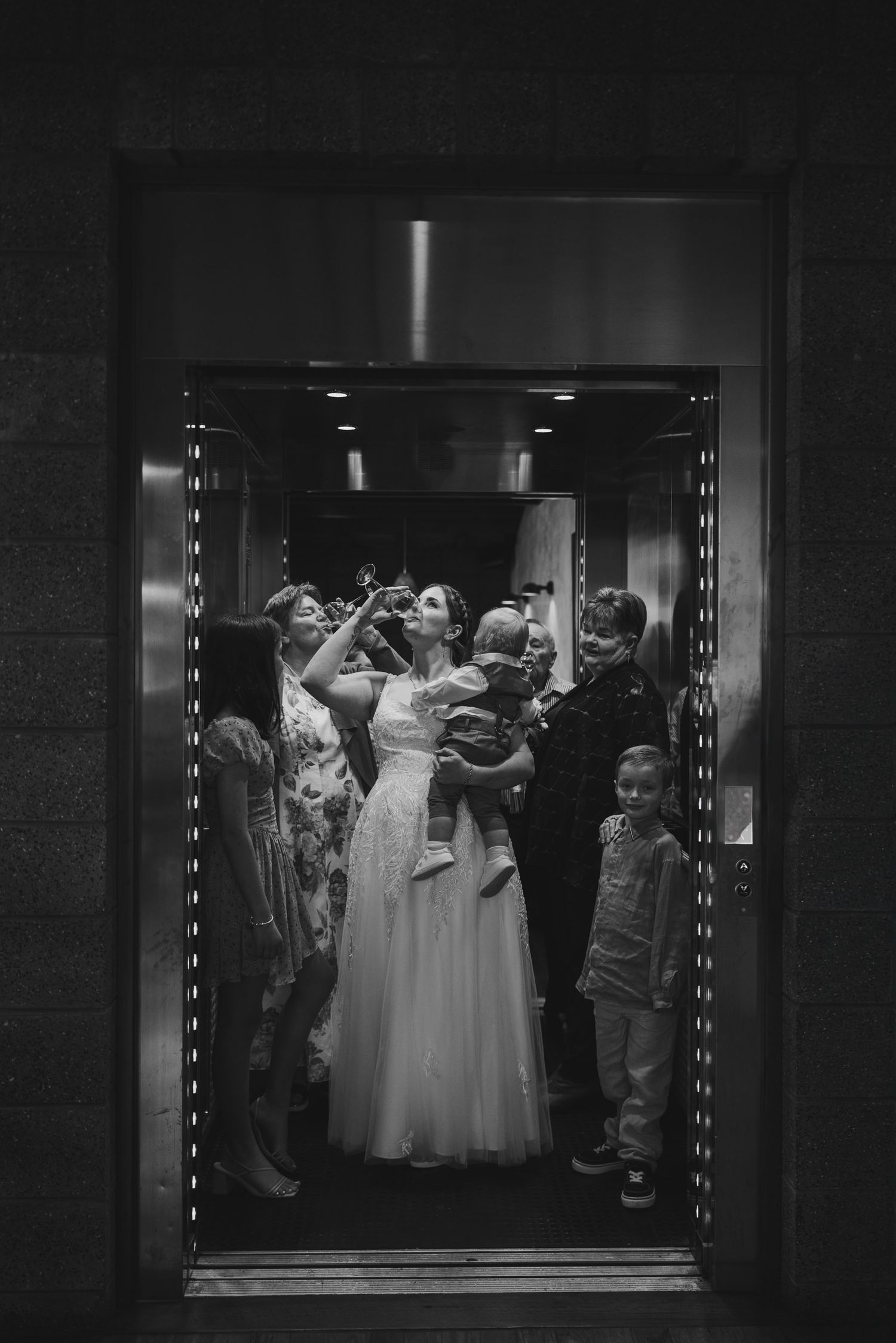 Black and white photo of a bride in a white gown surrounded by family in a crowded elevator.