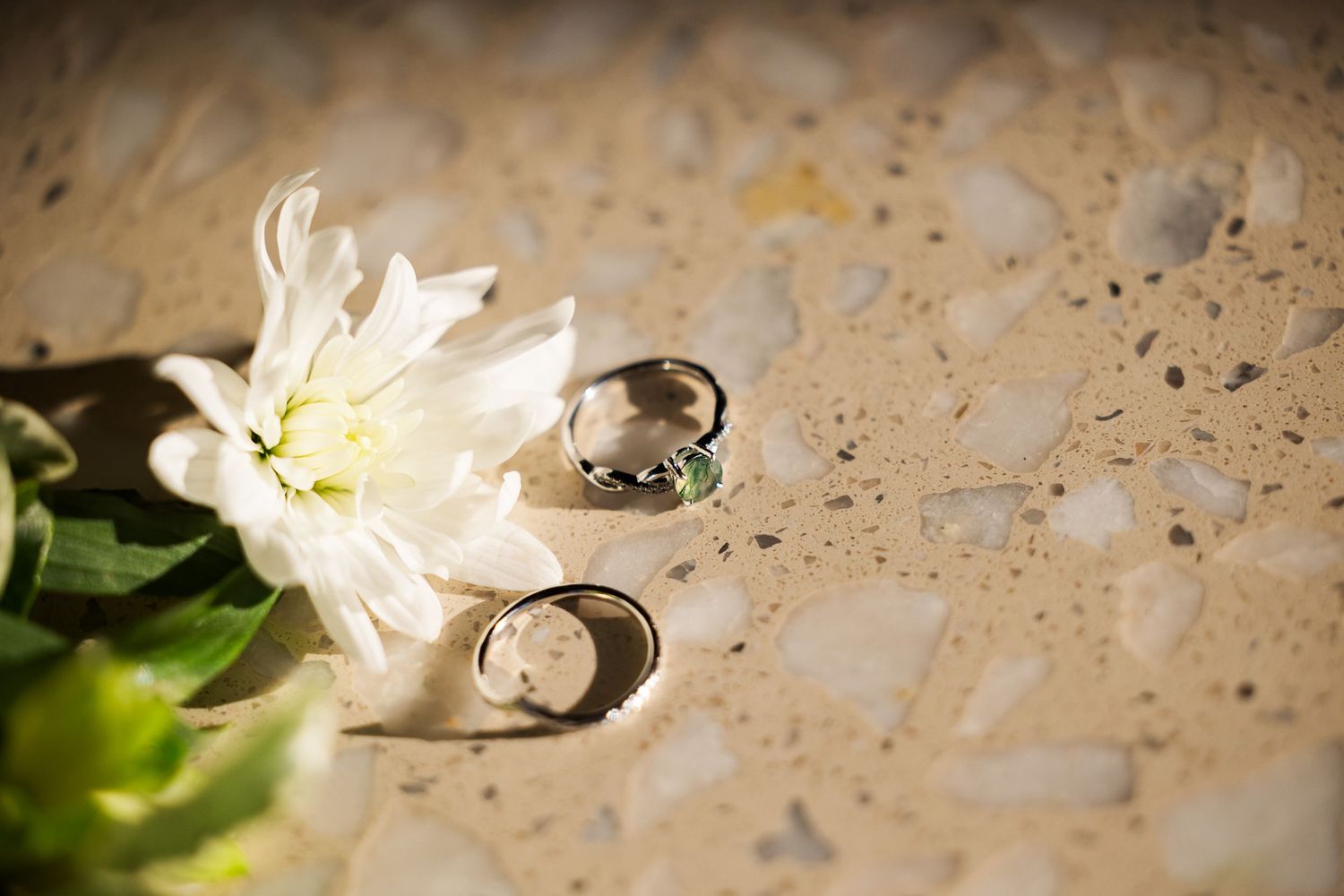 Two wedding rings and a white flower on a sandy textured surface, symbolizing love and commitment.