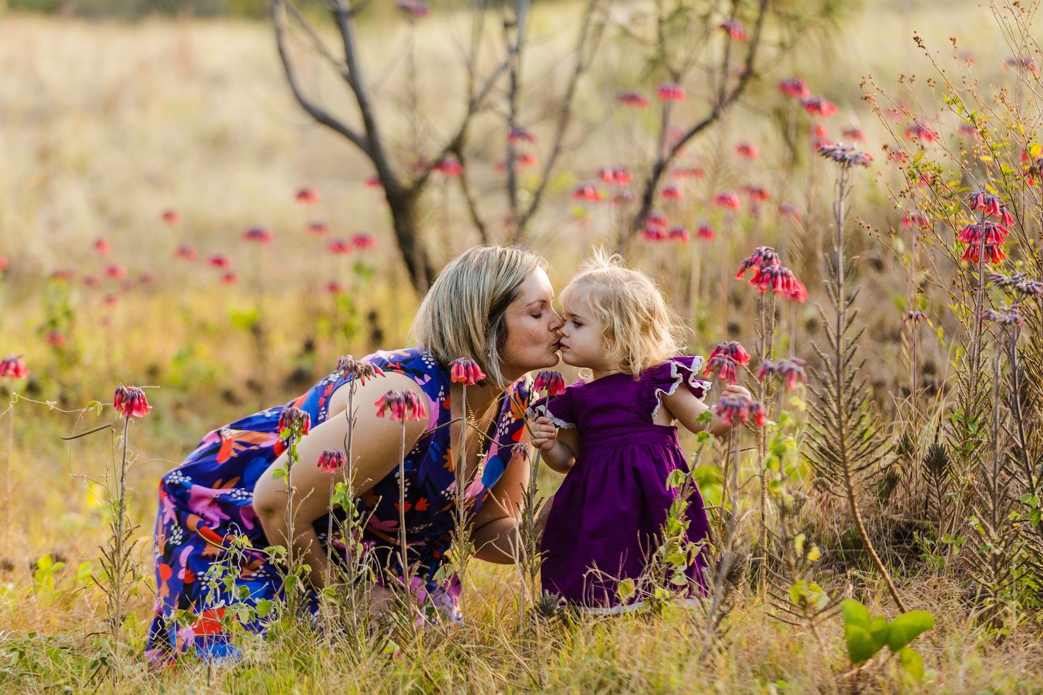 A mother and young daughter share a tender kiss outdoors in a wildflower field, wearing colorful dresses.