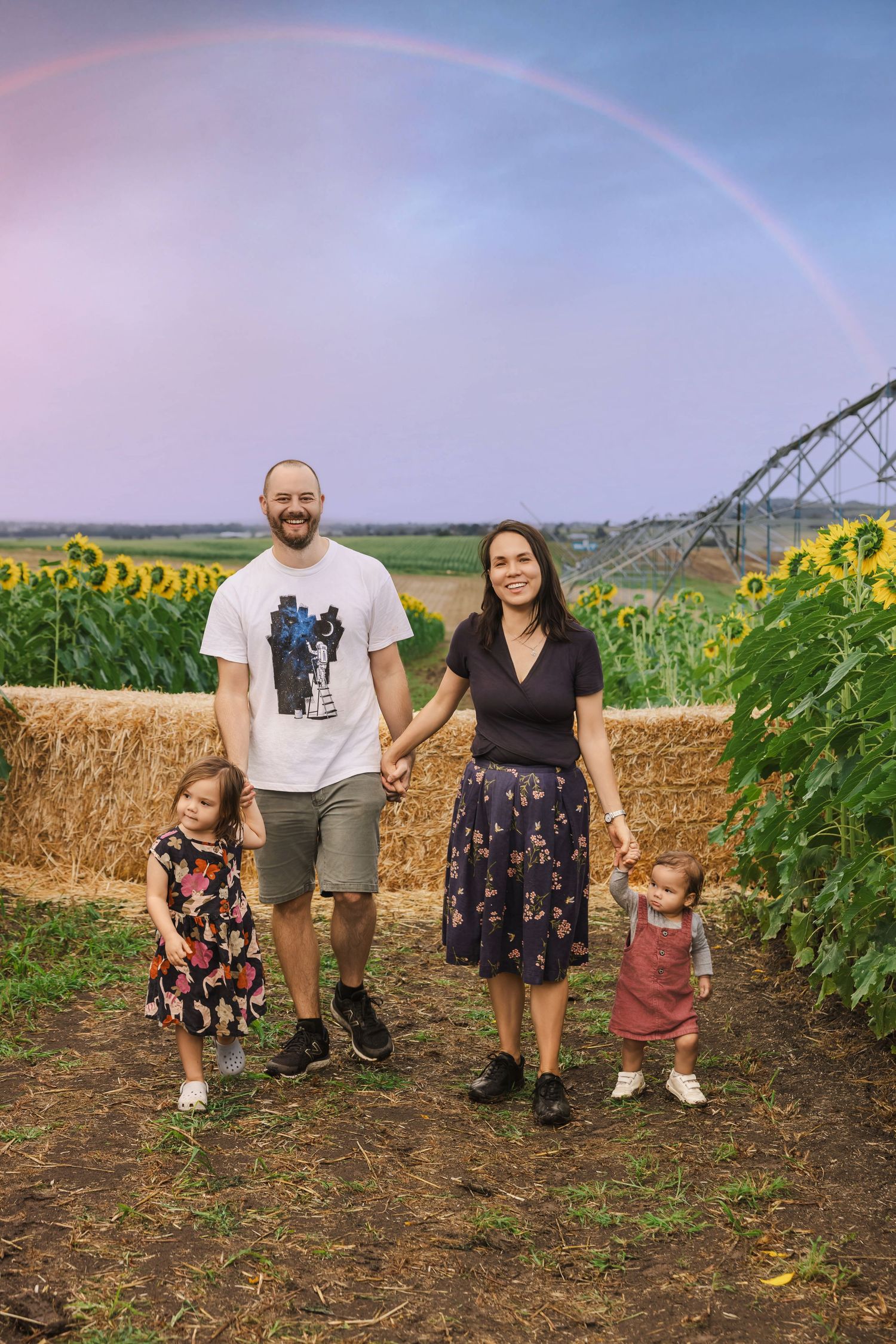 Smiling family of four holding hands near hay bales in a sunflower field with rainbow backdrop.