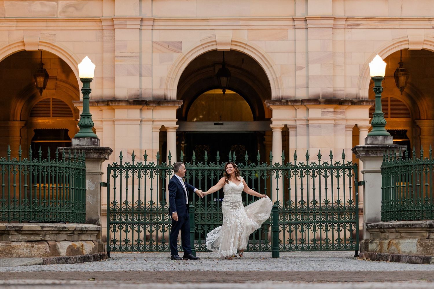 A couple in wedding attire poses for photos in front of an ornate stone building with arched columns and iron gates.