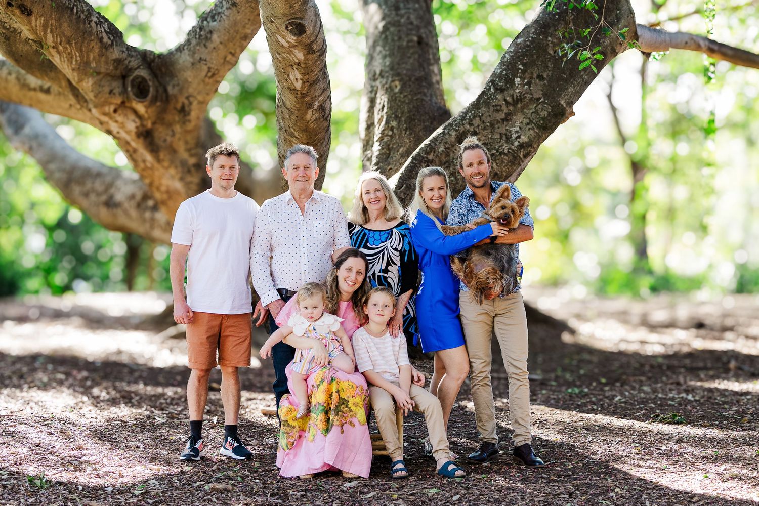 A group stands together under a large tree in a park setting with a small dog.