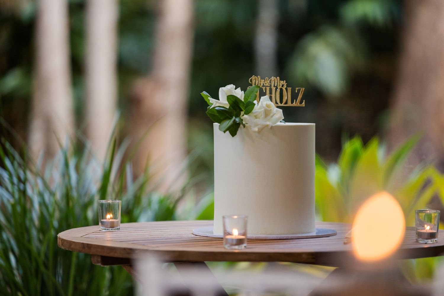 Elegant white wedding cake decorated with greenery and gold accents sits on wooden table surrounded by tropical plants.
