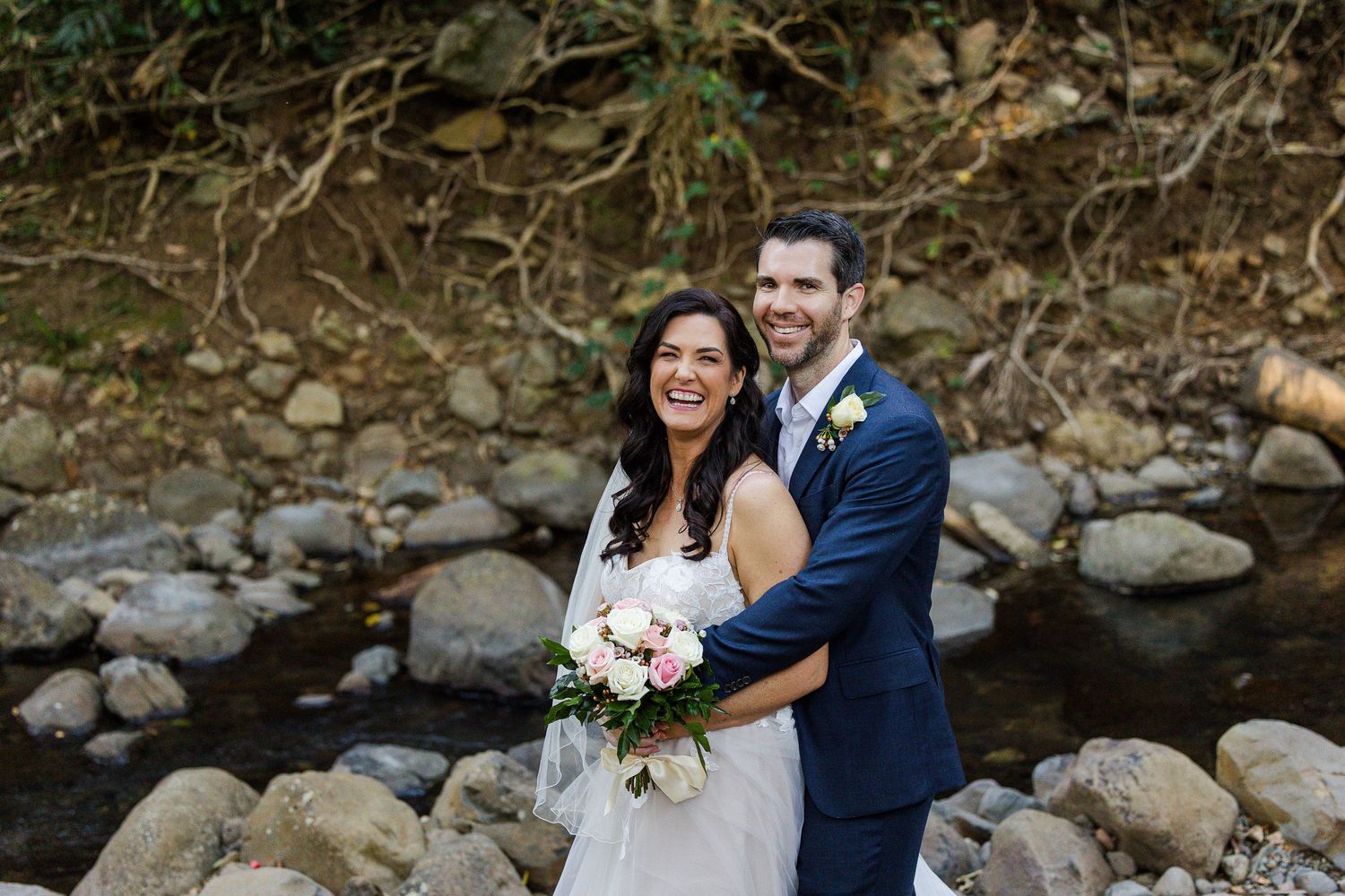 Wedding couple sharing joyful moments together by a rocky stream in an outdoor setting.