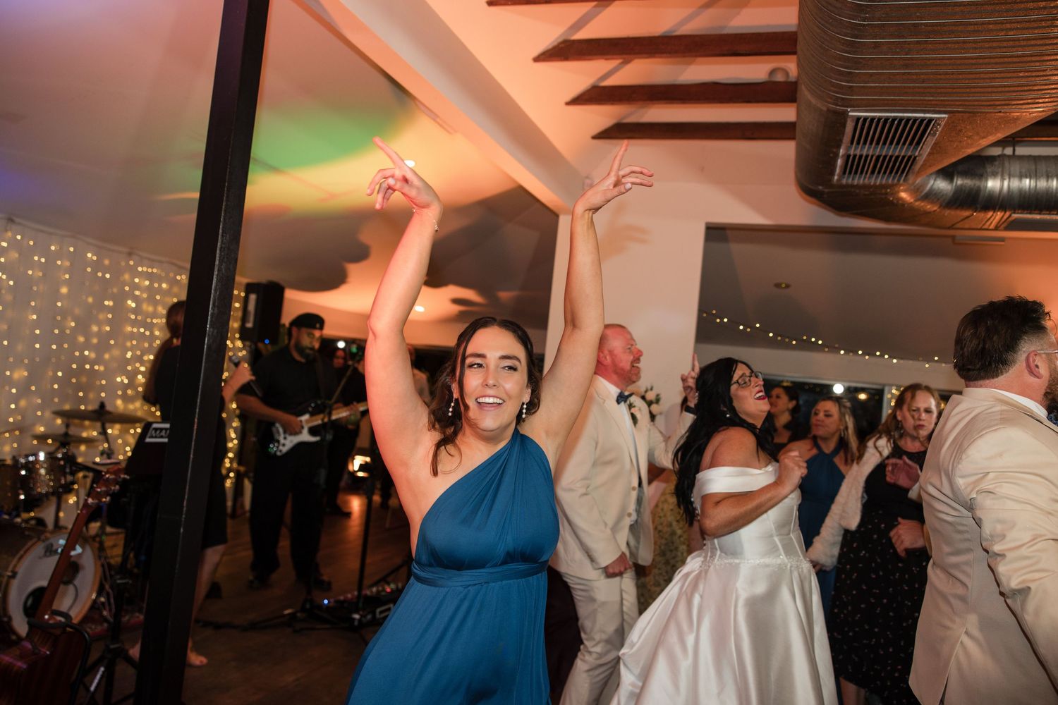 Bridesmaid in blue dress dancing with arms raised at a wedding reception with fairy lights.