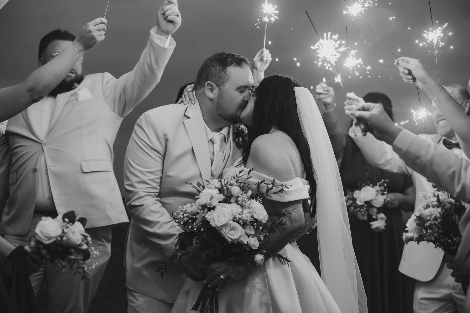 Black and white wedding photo of bride and groom kissing under sparklers held by guests.